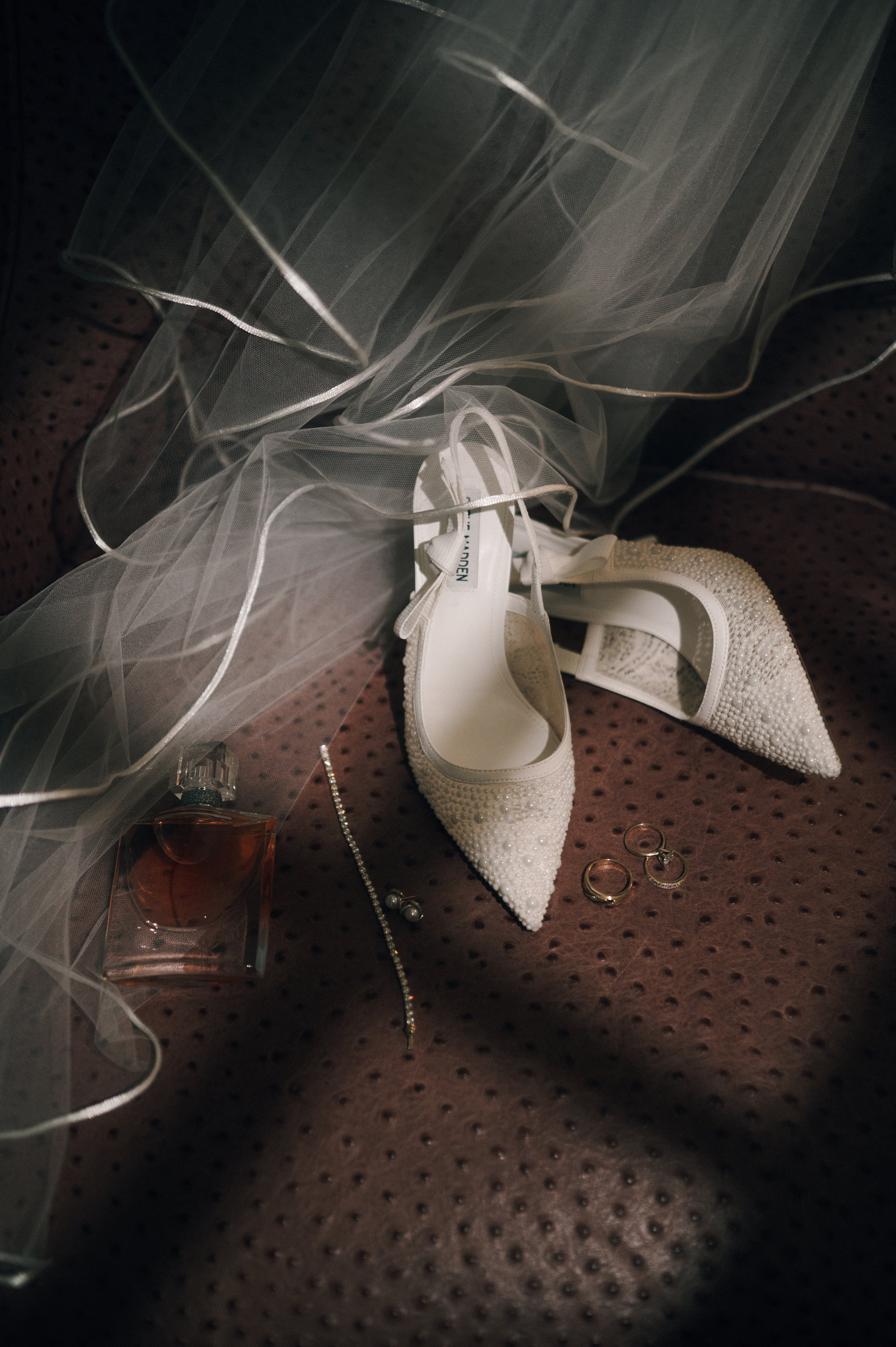 A pair of white bridal shoes decorated with pearls, placed on a maroon polka-dotted fabric. Surrounding the shoes are rings, earrings, a necklace, a perfume bottle, and a bridal veil.