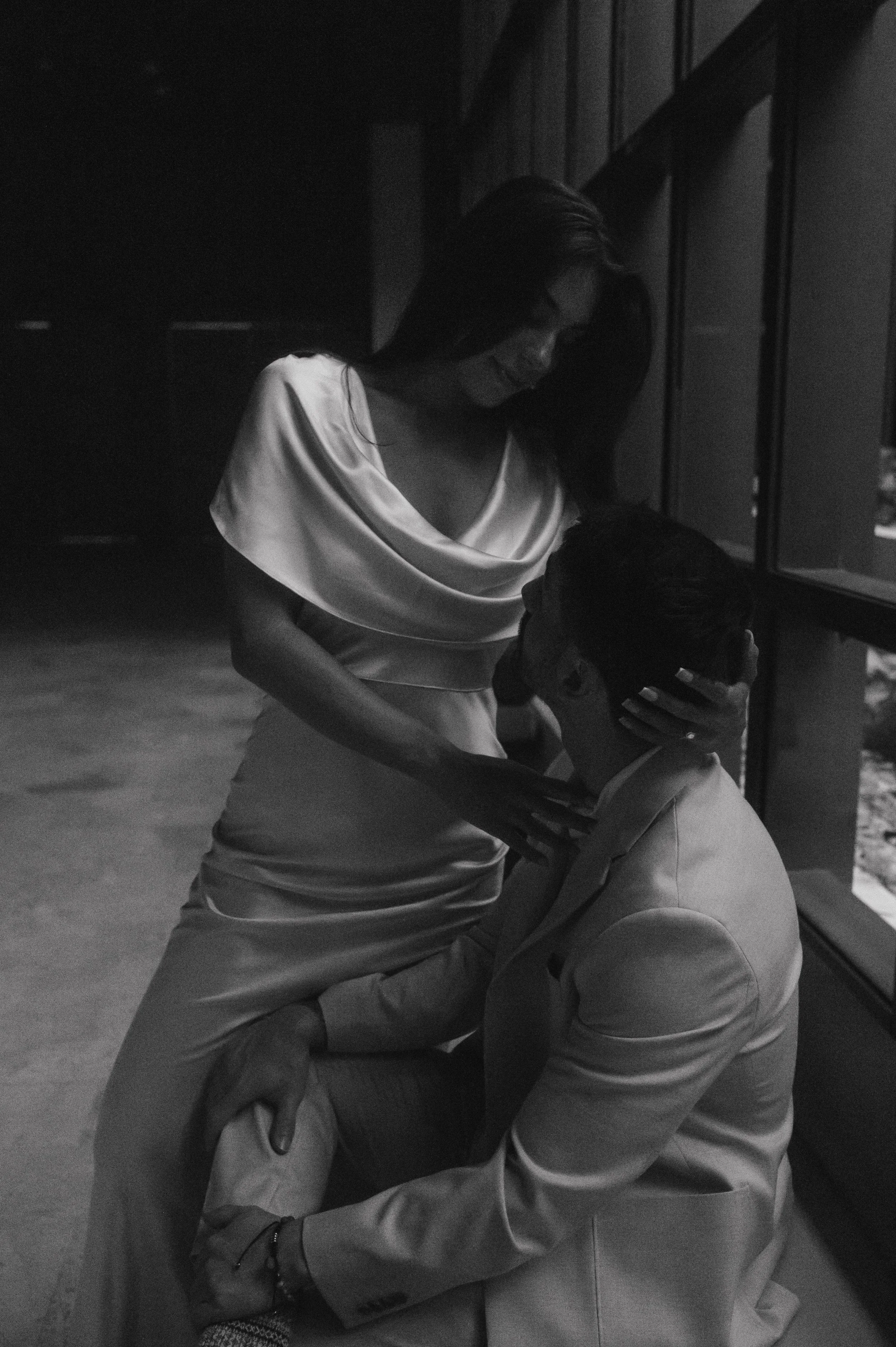 A woman in a satin dress is smiling as she interacts with a seated man, in an indoor setting with dark walls and large windows.