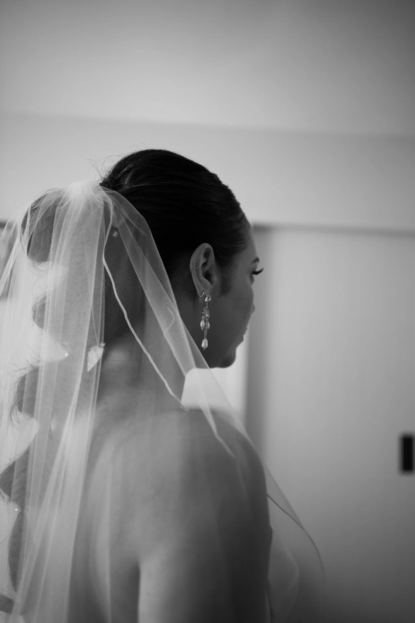 Black and white photo of a woman wearing a bridal headpiece and veil, with earrings, seen from the side profile.