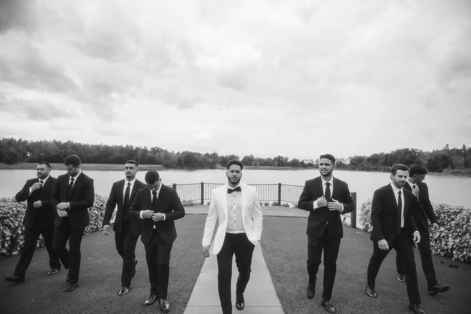 Group of seven men walking outdoors near a lake, dressed formally in suits and tuxedos, with one man in a white tuxedo jacket and black bow tie leading, overcast sky.