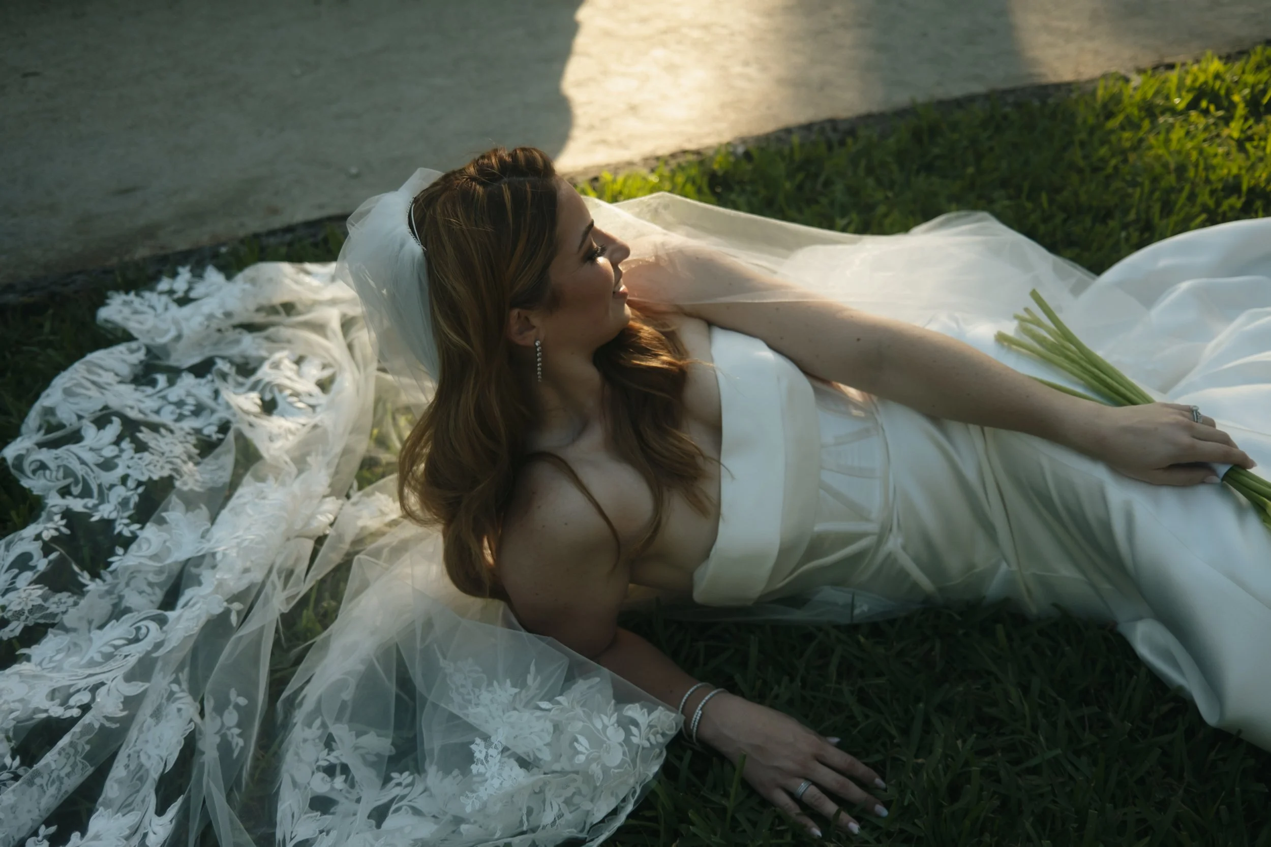 A woman in a wedding dress laying on grass beside a sidewalk, holding a bouquet of flowers, with her eyes closed and smiling.