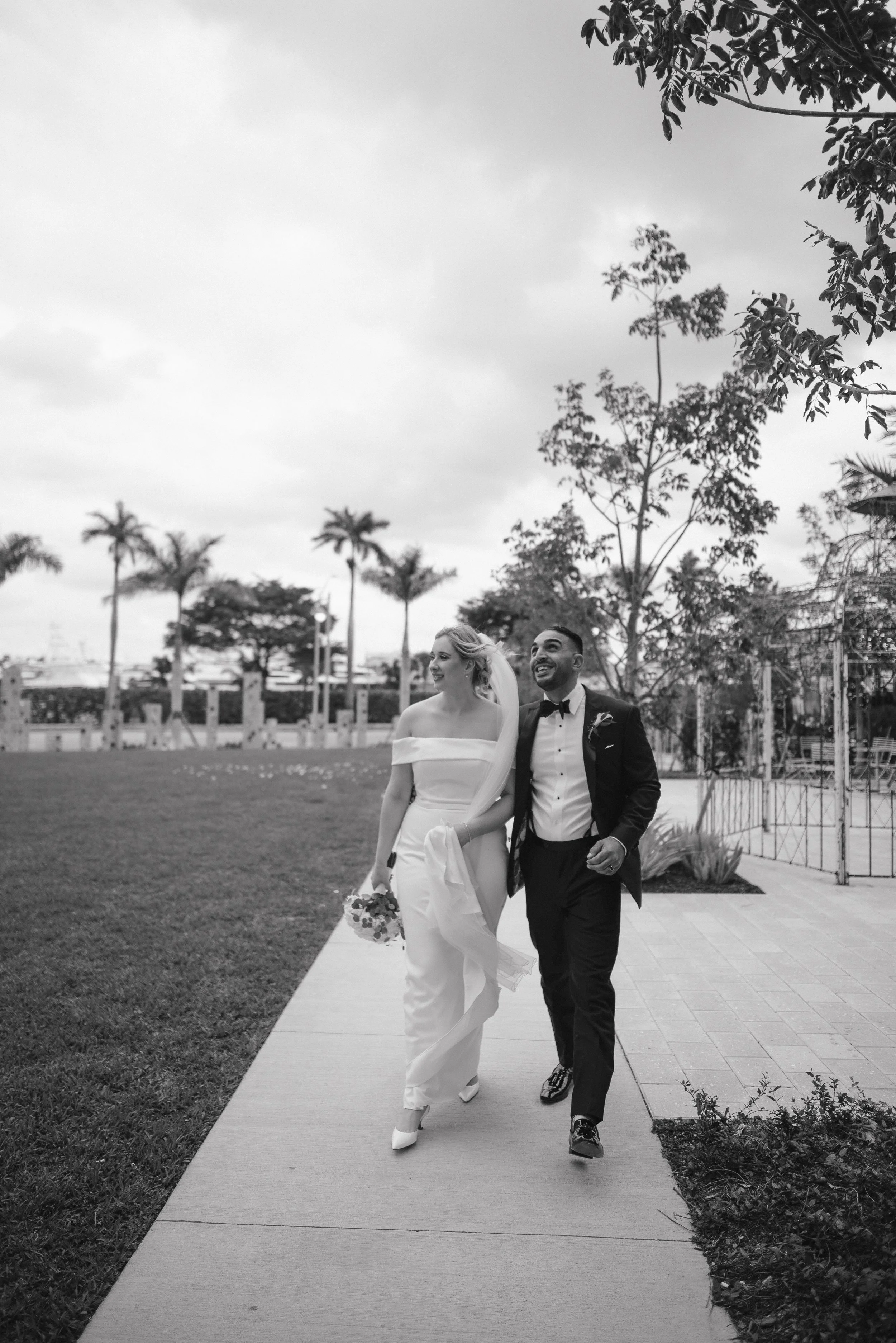 A black and white photo of a newlywed couple walking outdoors, the bride in a white off-the-shoulder gown with a veil and holding a bouquet, the groom in a tuxedo with a bow tie, both smiling and looking happy.