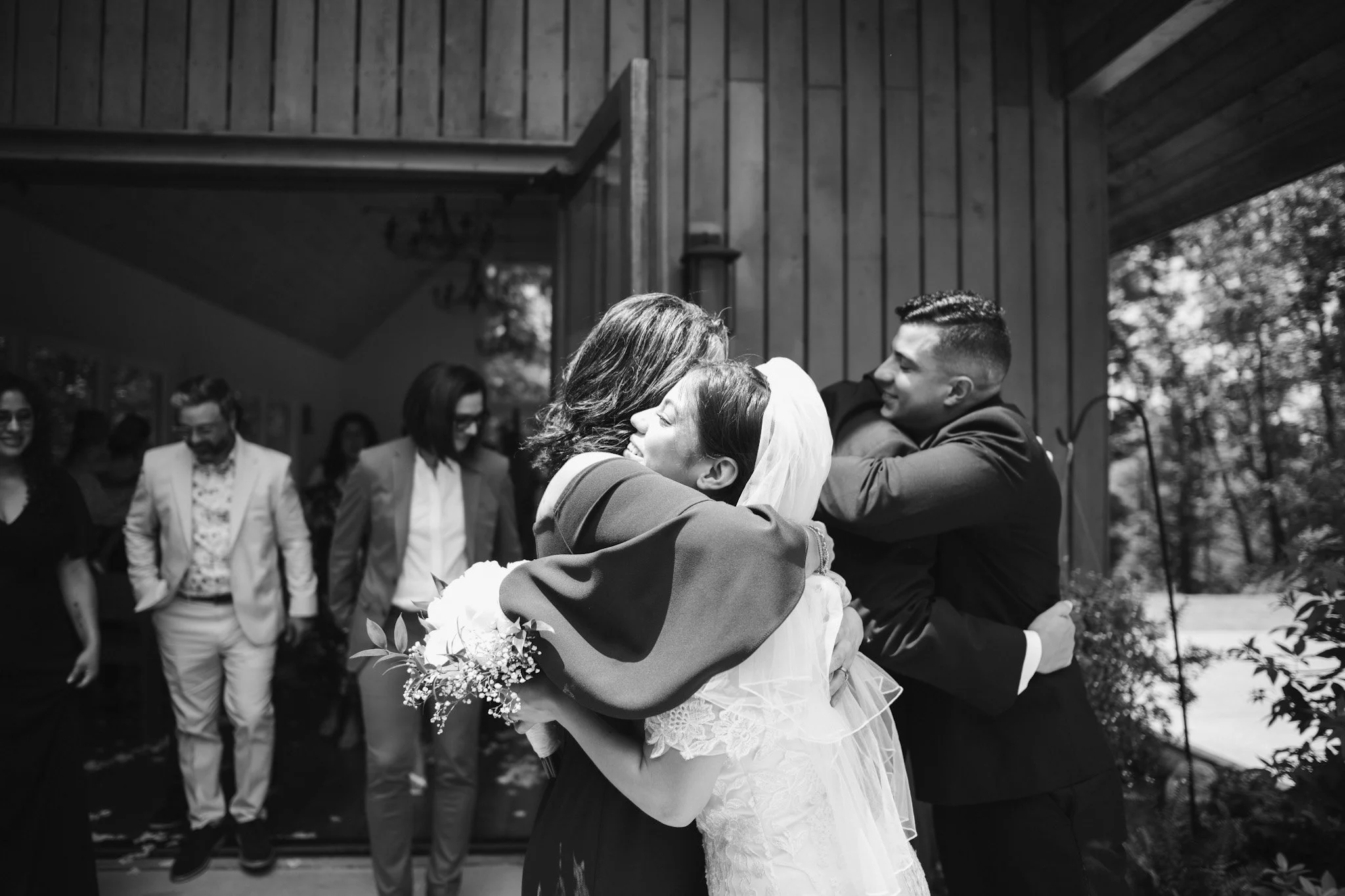 A group of people at a wedding reception, with a bride and groom embracing and smiling, while others in the background are dressed formally and smiling.