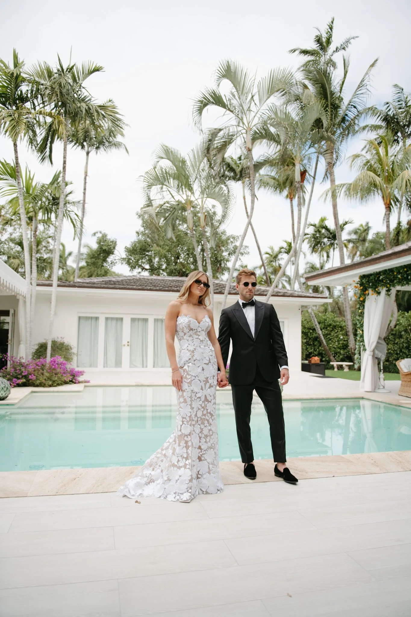 Couple walking hand in hand by a swimming pool, surrounded by palm trees and white buildings, during a sunny day.