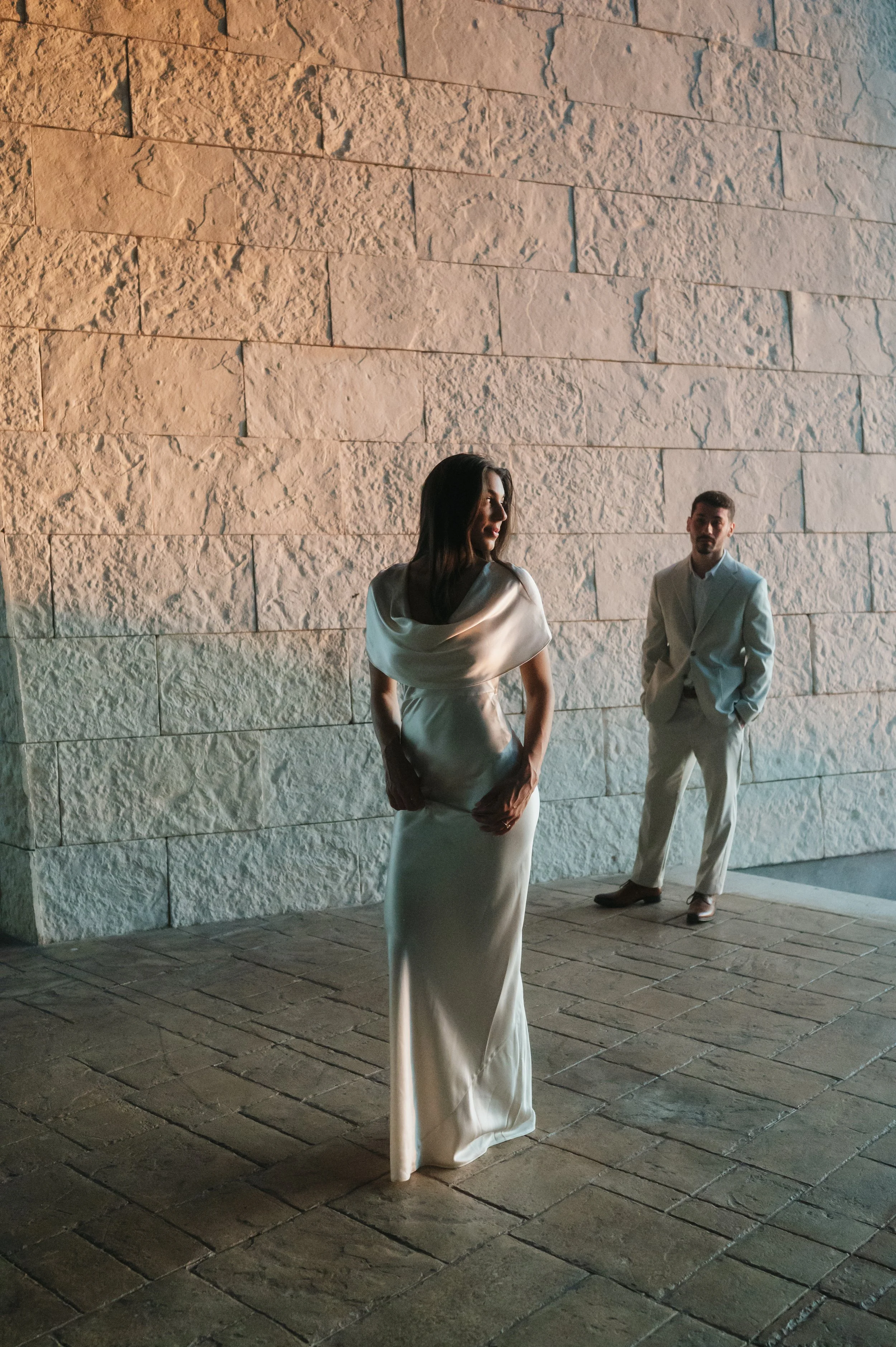 A woman in a white gown standing in front of a stone wall, with a man in a matching white suit standing in the background, outdoors at night.