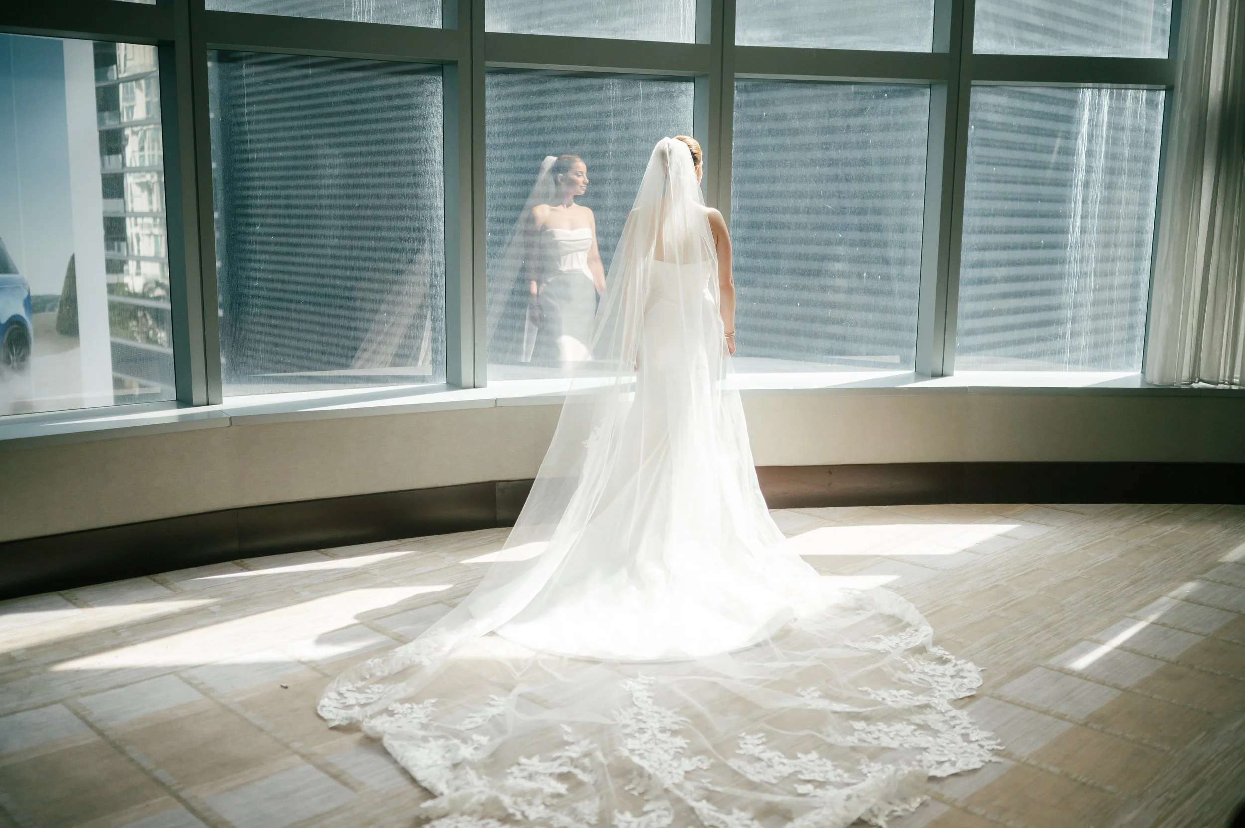 A bride in a white wedding dress and veil standing by large windows, gazing at her reflection in the glass.