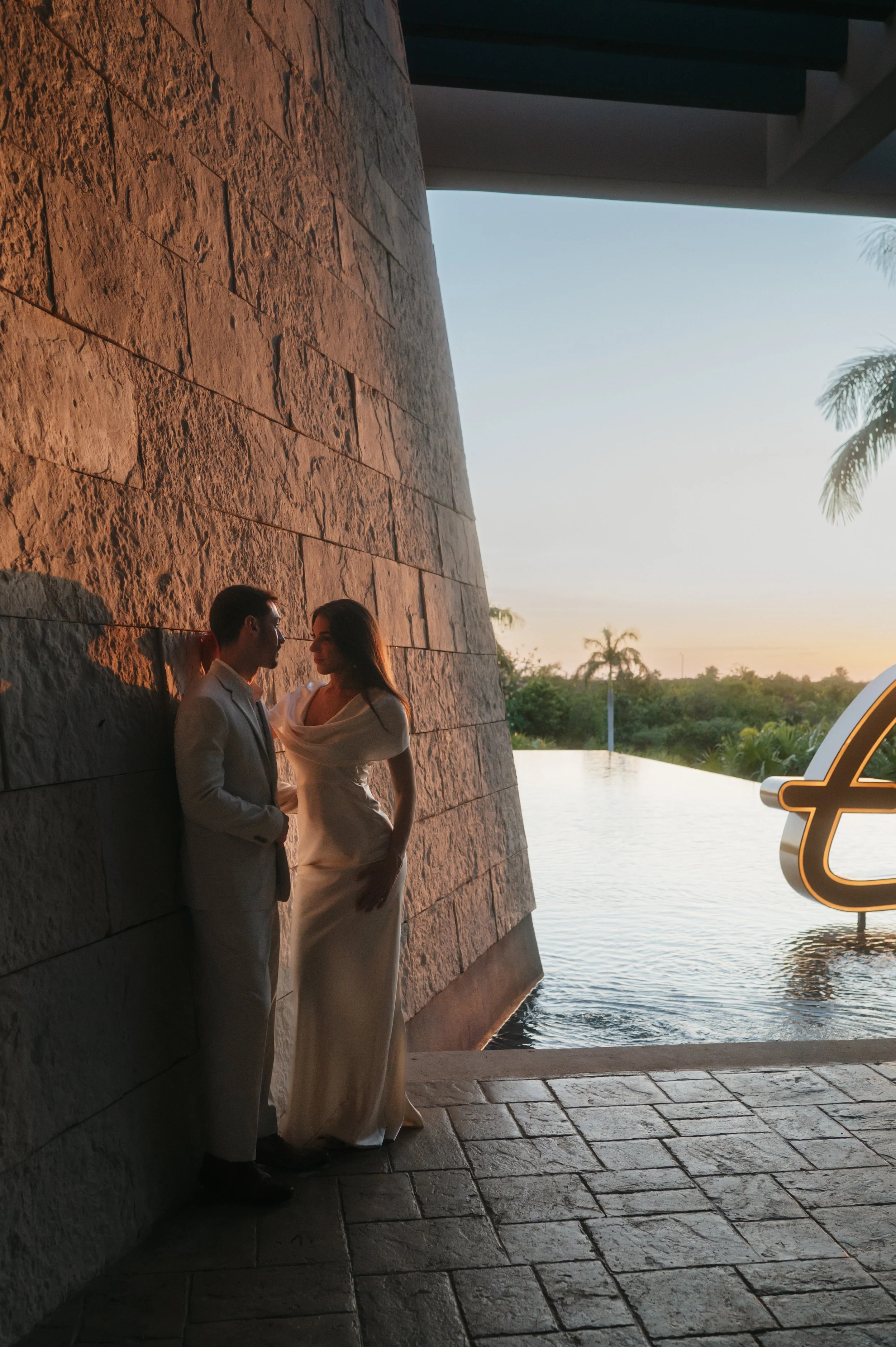 A man and woman dressed in formal attire standing close to each other against a textured stone wall during sunset by a body of water, with palm trees in the background.