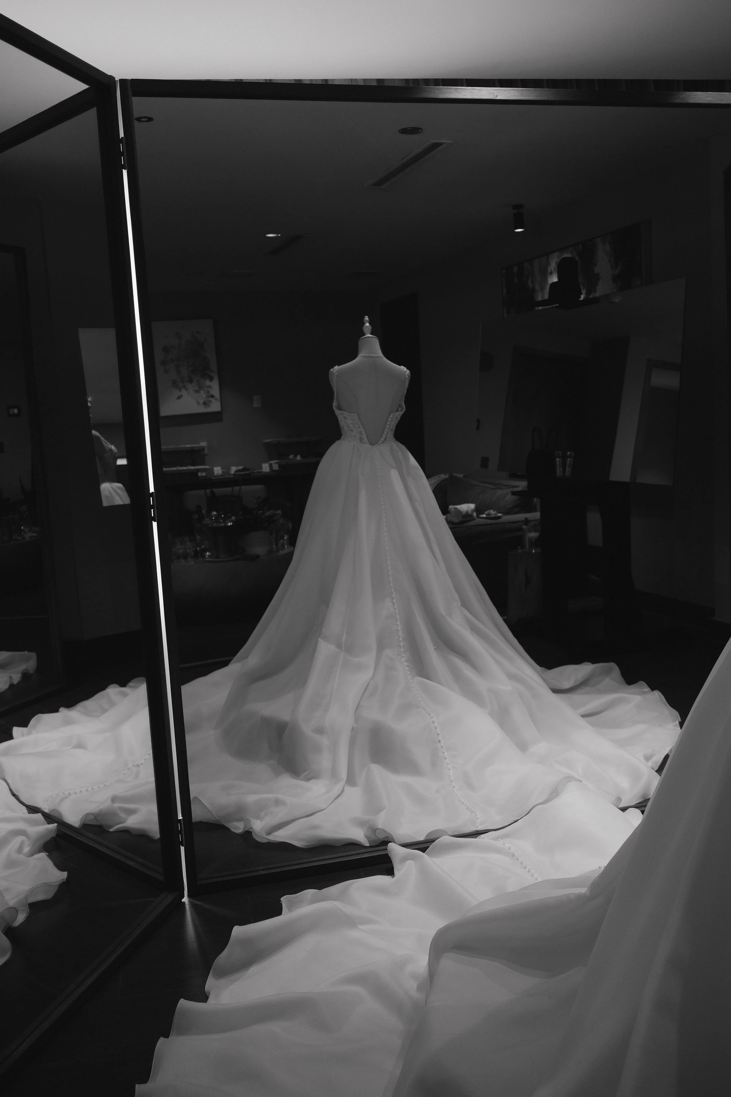 A wedding dress displayed on a mannequin inside a glass enclosure, with fabric pooling on the floor around it, in a room with artwork and furniture.