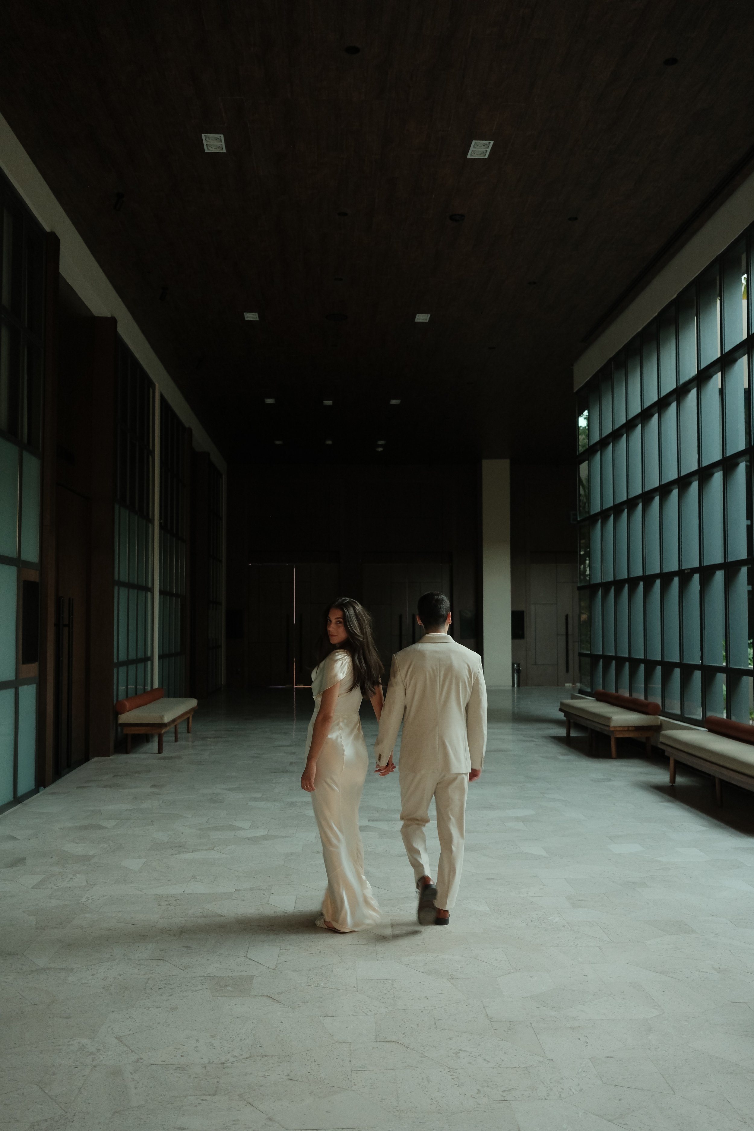 A woman and a man dressed in formal white attire walking hand in hand through a spacious, modern interior with large windows, benches, and a wooden ceiling.
