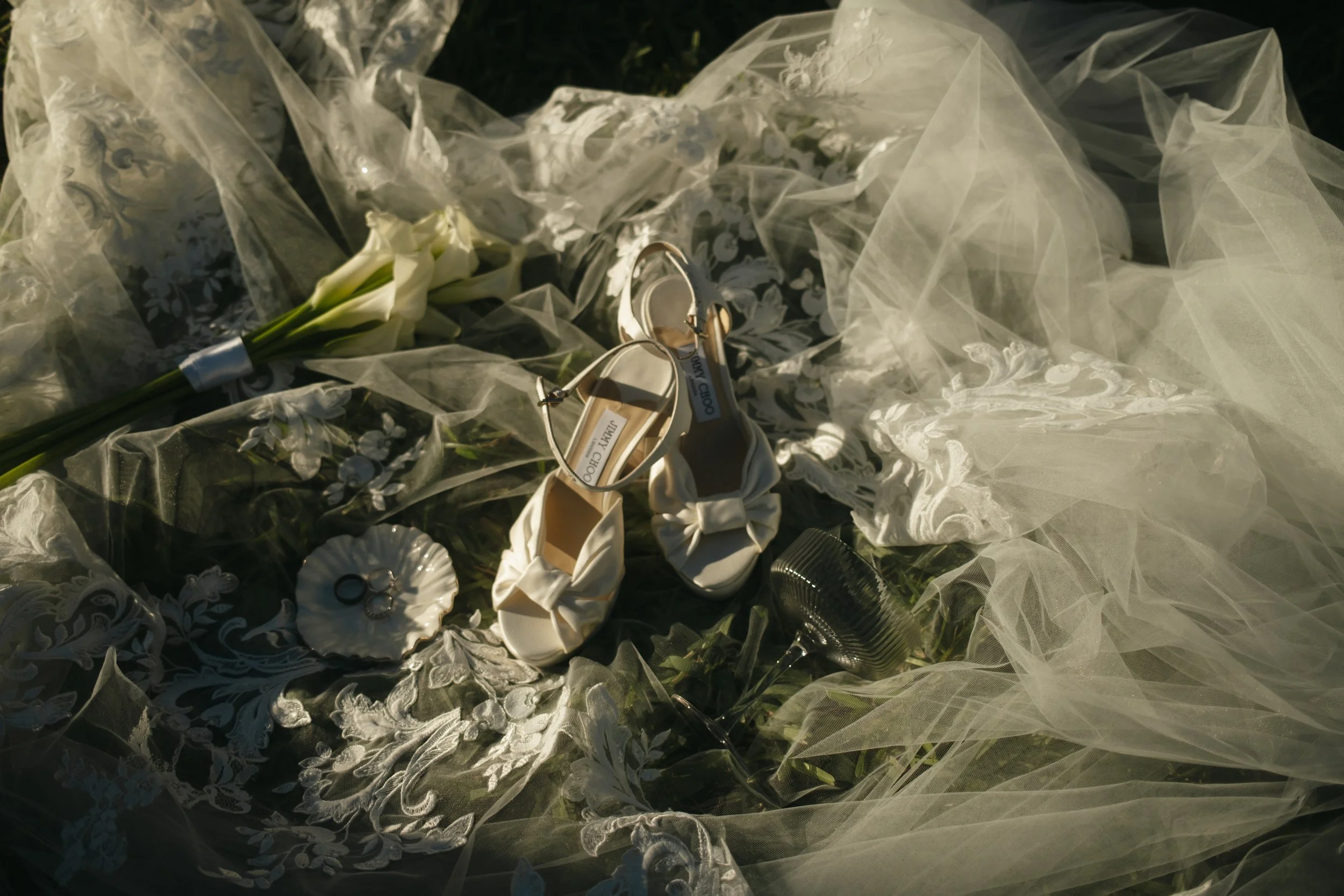 A wedding veil, white shoes, a bouquet of white lilies, and jewelry are arranged on lace fabric and tulle in a setting that appears to be at an outdoor wedding or event.