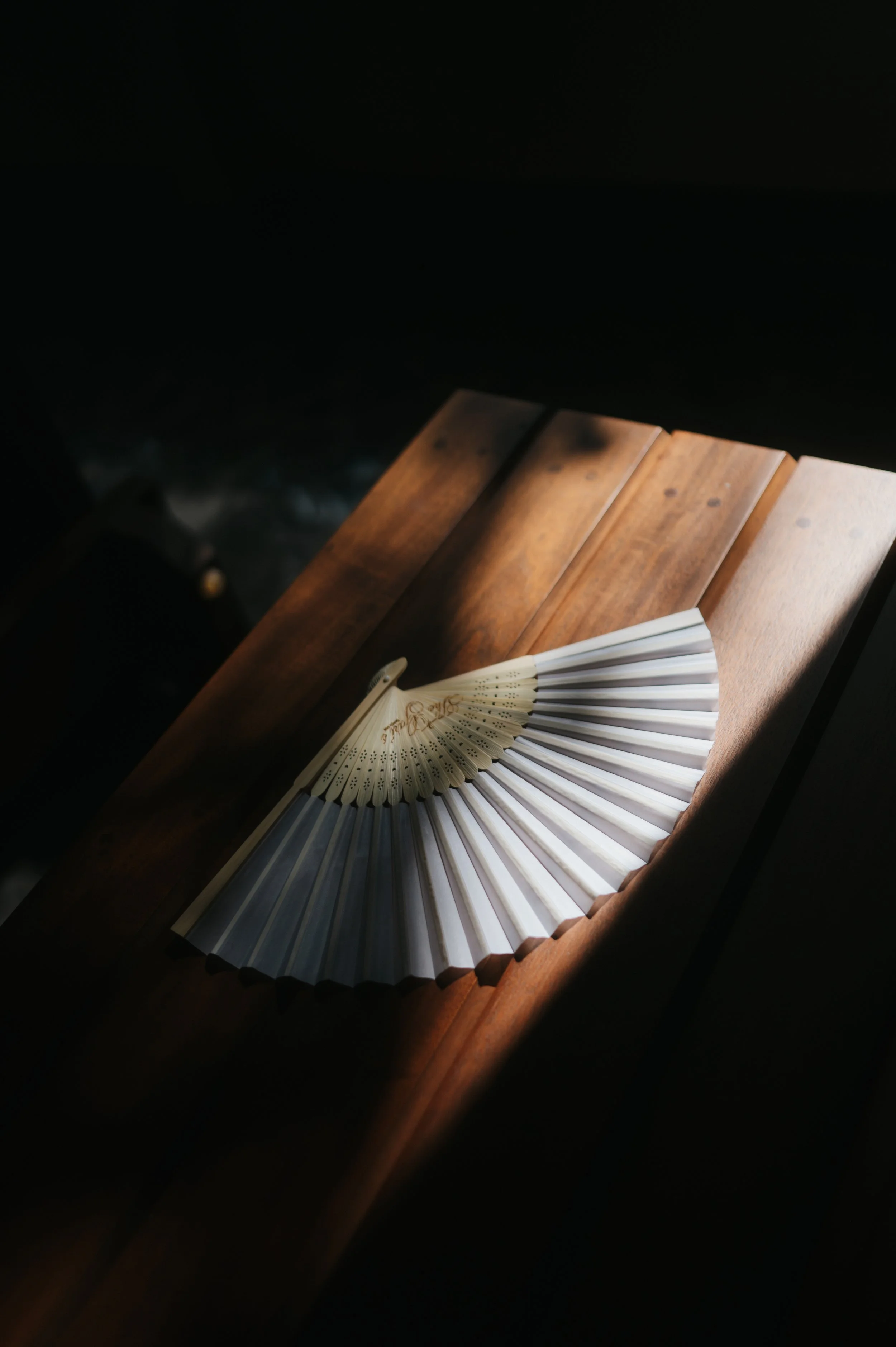 A wooden table with a partially opened white folding fan resting on it, with the dark background obscuring the surrounding area.