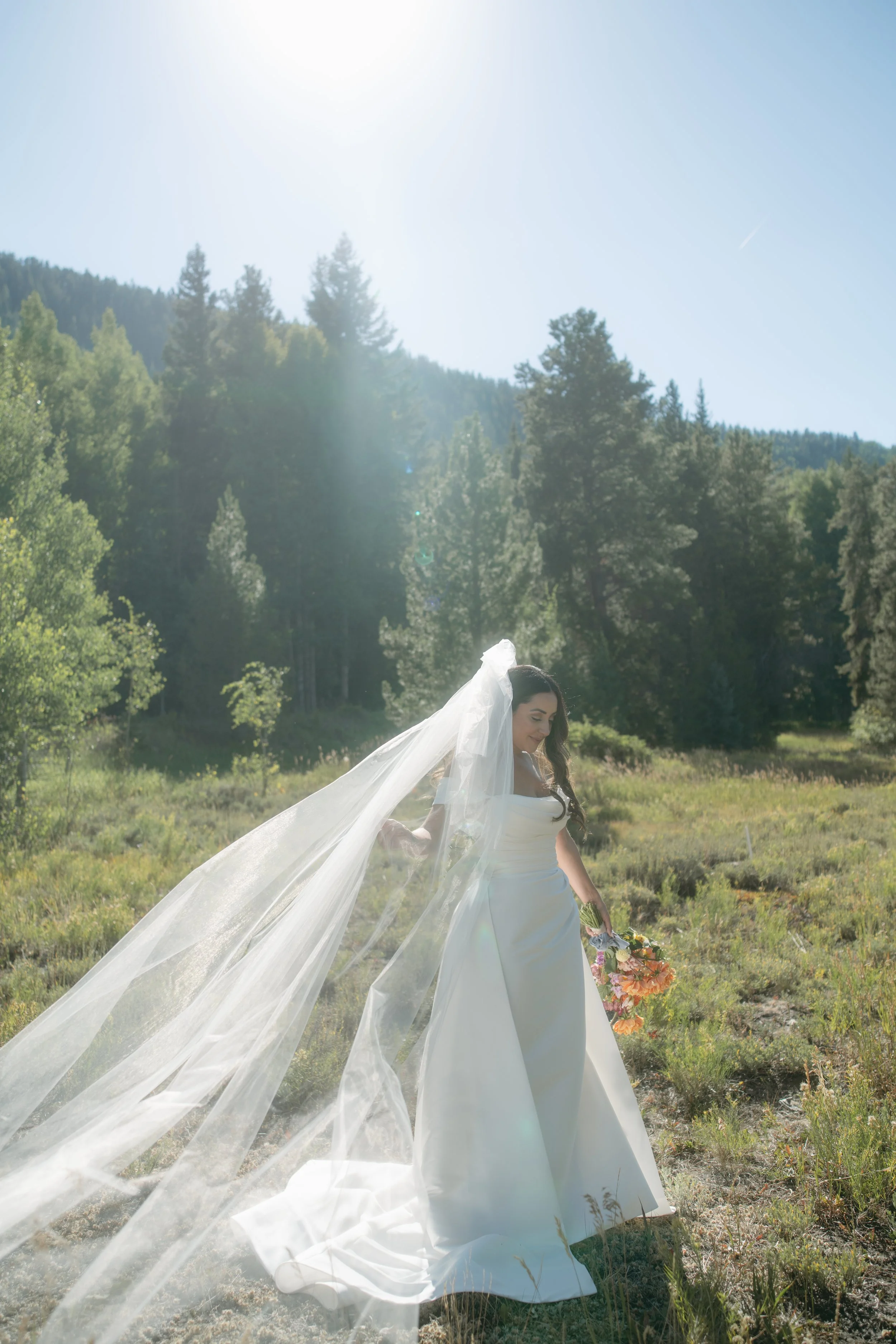 Bride in a white wedding dress and veil standing outdoors in a grassy field with trees and mountains in the background, holding a bouquet of flowers.