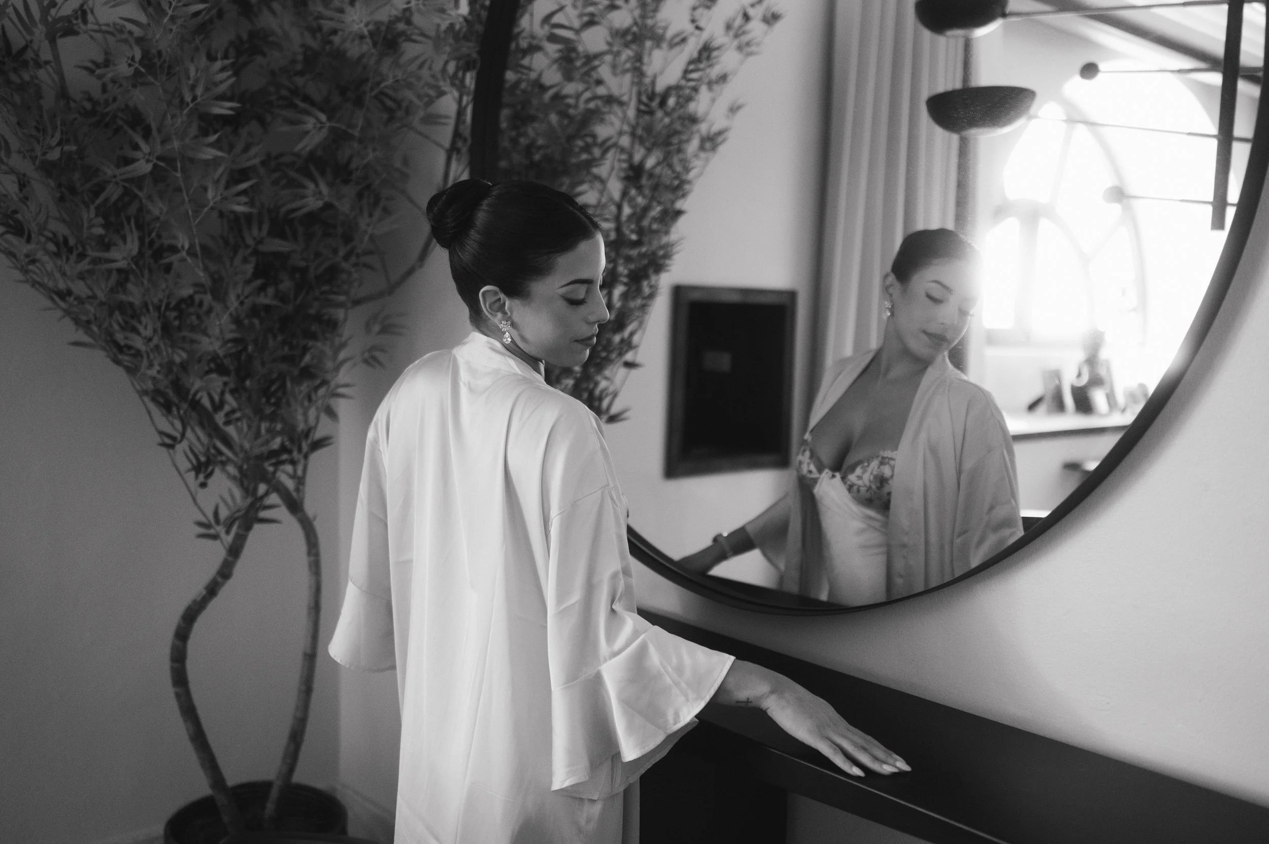 A woman with dark hair styled in an elegant bun, wearing a satin robe and earrings, looking at her reflection in a mirror. The scene is in a well-lit room with a potted plant in the foreground.