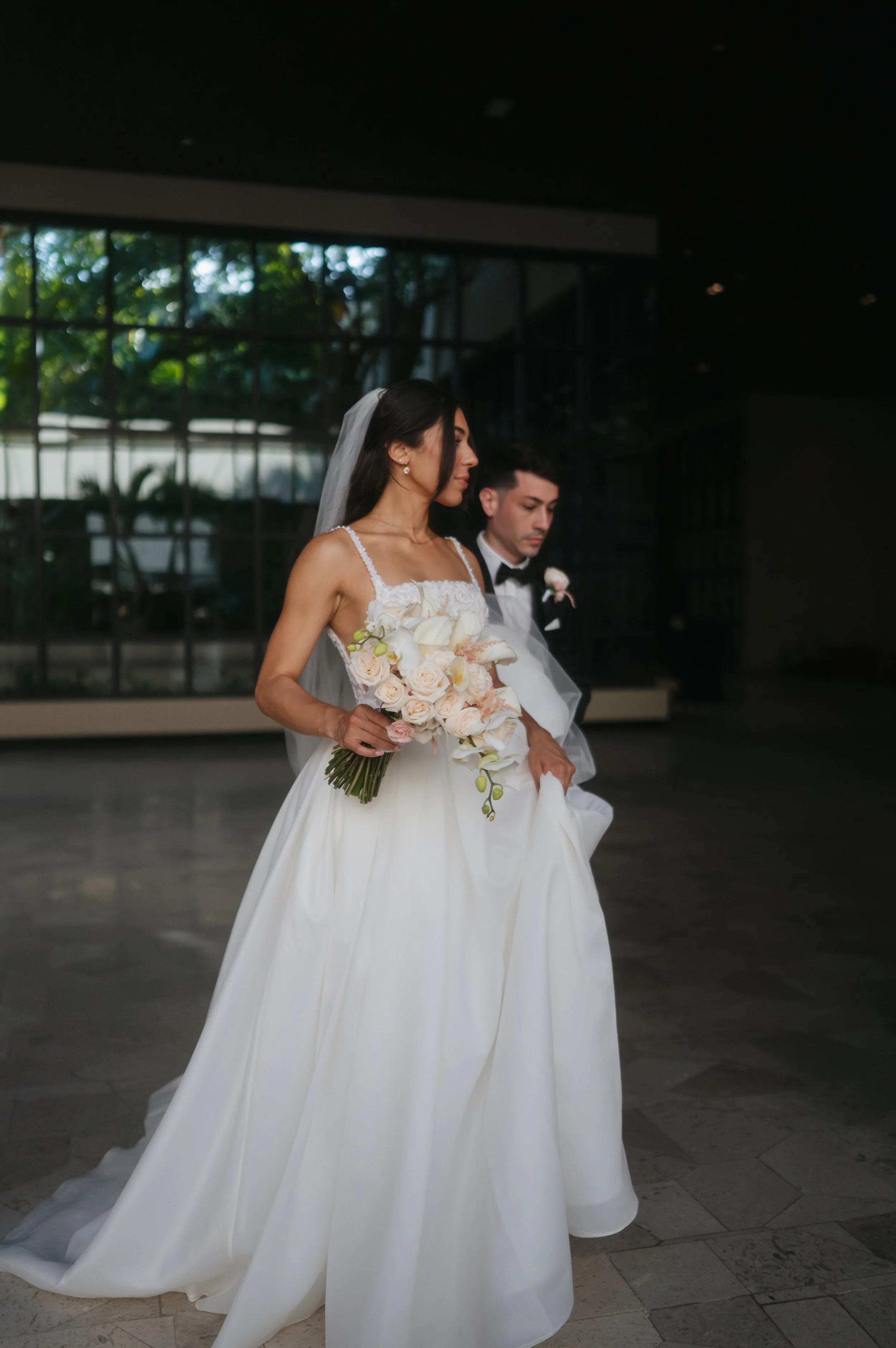 Bride in a white wedding gown holding a bouquet of white and soft pink flowers, standing indoors with a man in a tuxedo behind her, against a background of large windows with trees outside.