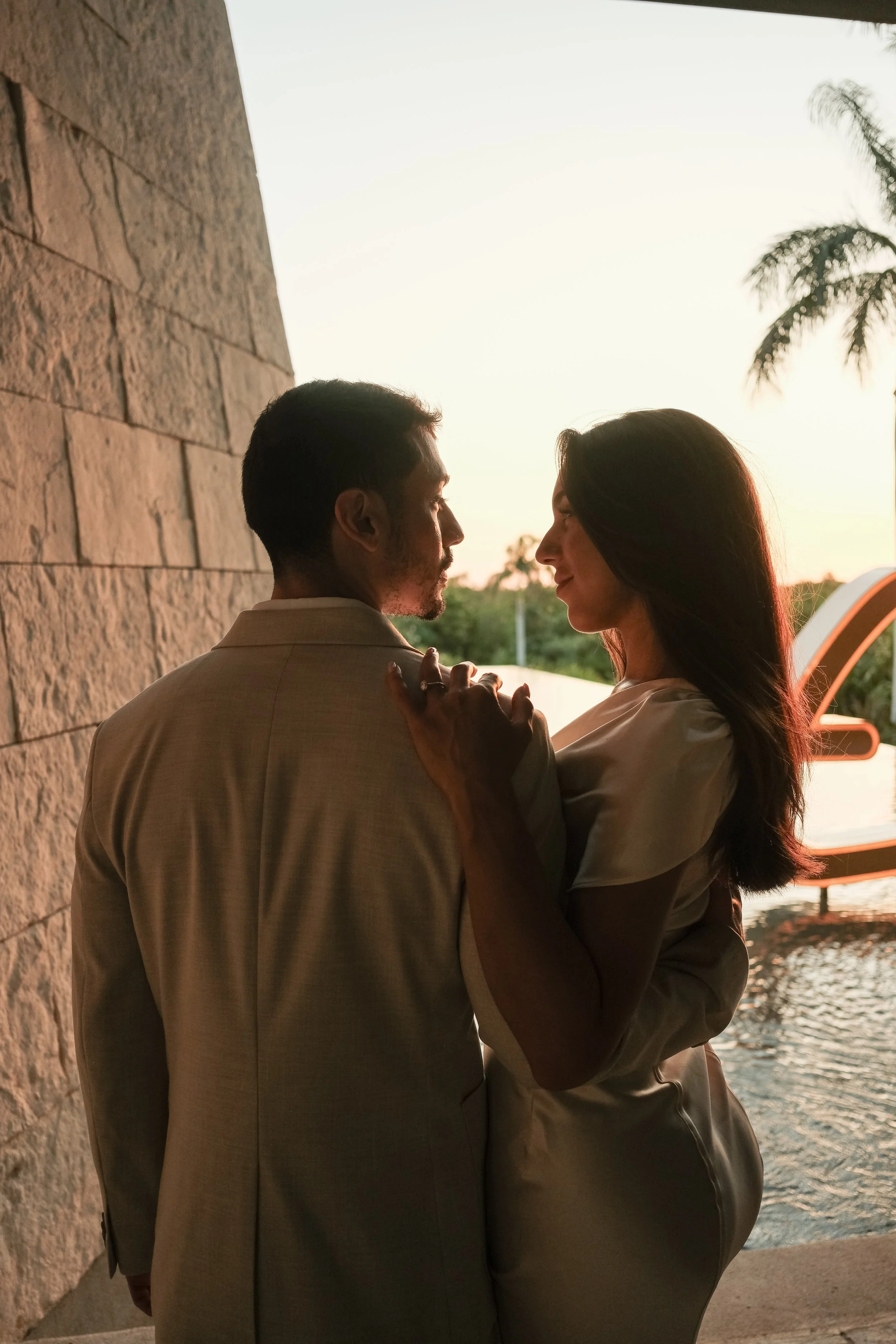 A man and woman dancing closely by a waterfront during sunset, with trees and a boat in the background.