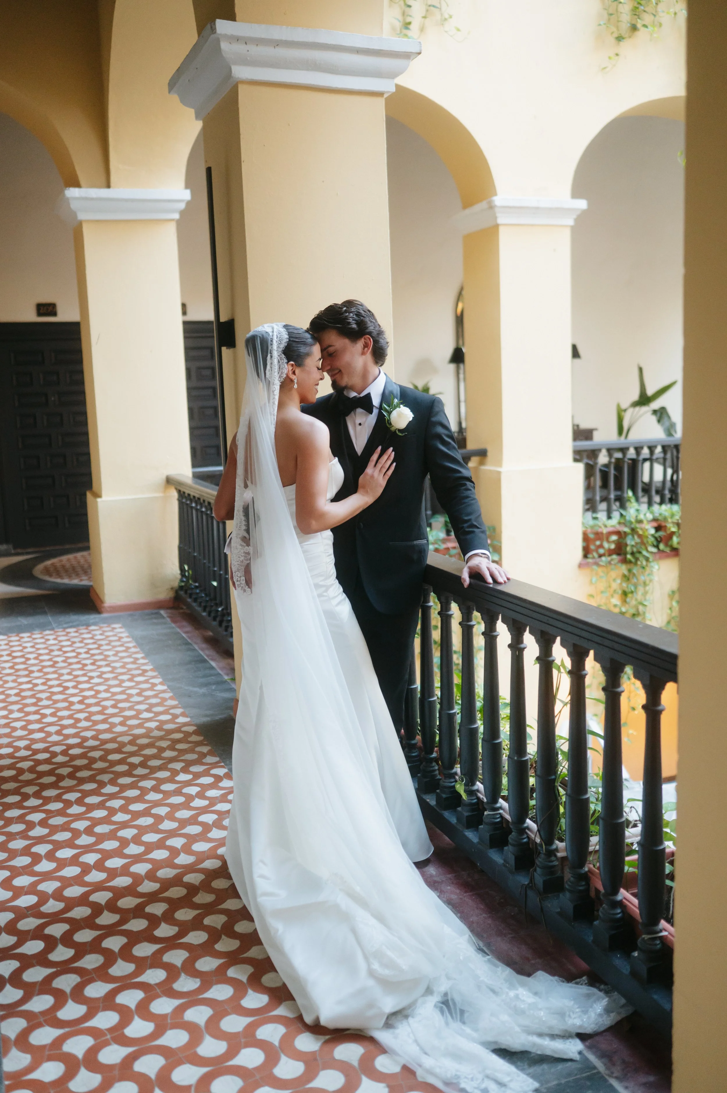 A bride and groom standing close together on a balcony with arches, in an indoor or outdoor courtyard, embracing with their foreheads touching and eyes closed, dressed in wedding attire.