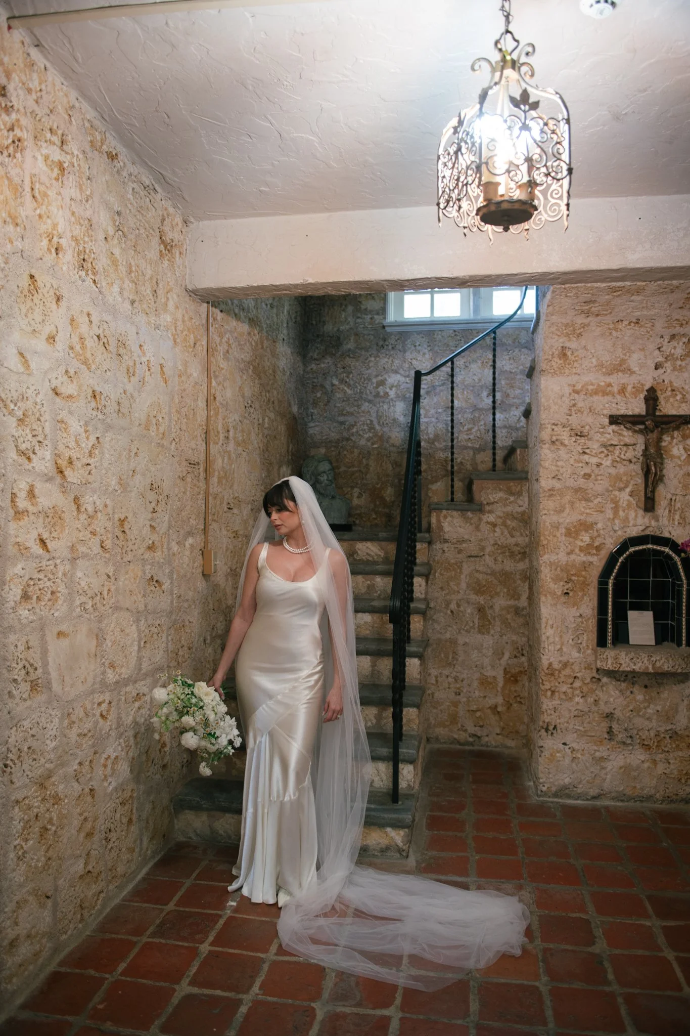 Bride in a satin wedding gown holding a bouquet in a rustic stone interior with a staircase and religious cross