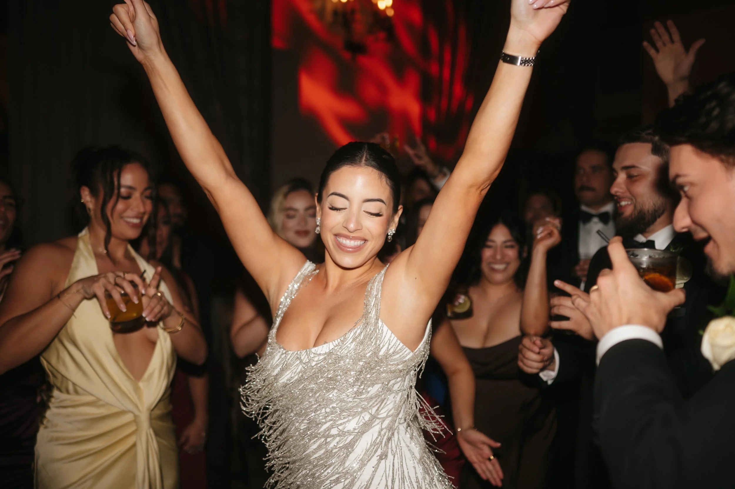 A woman in a silver, glittering dress dancing with her eyes closed and arms raised at a lively party with other guests enjoying drinks and smiling.
