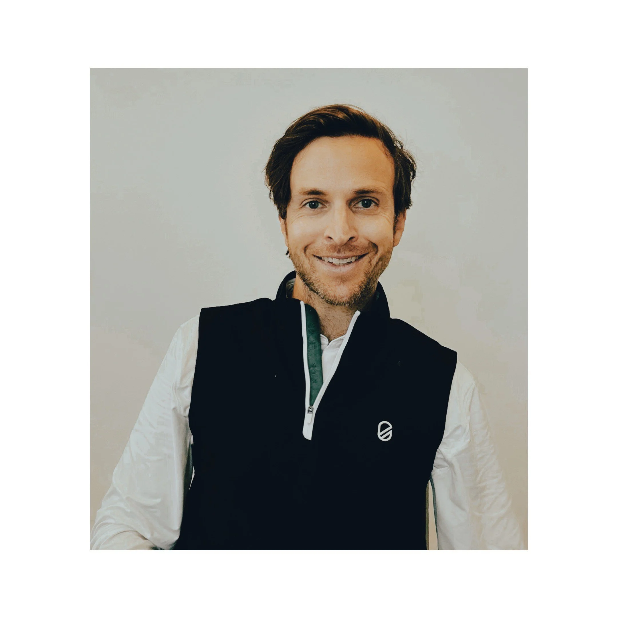 A smiling man with brown hair and blue eyes, wearing a white shirt and a black vest, standing against a plain white background.