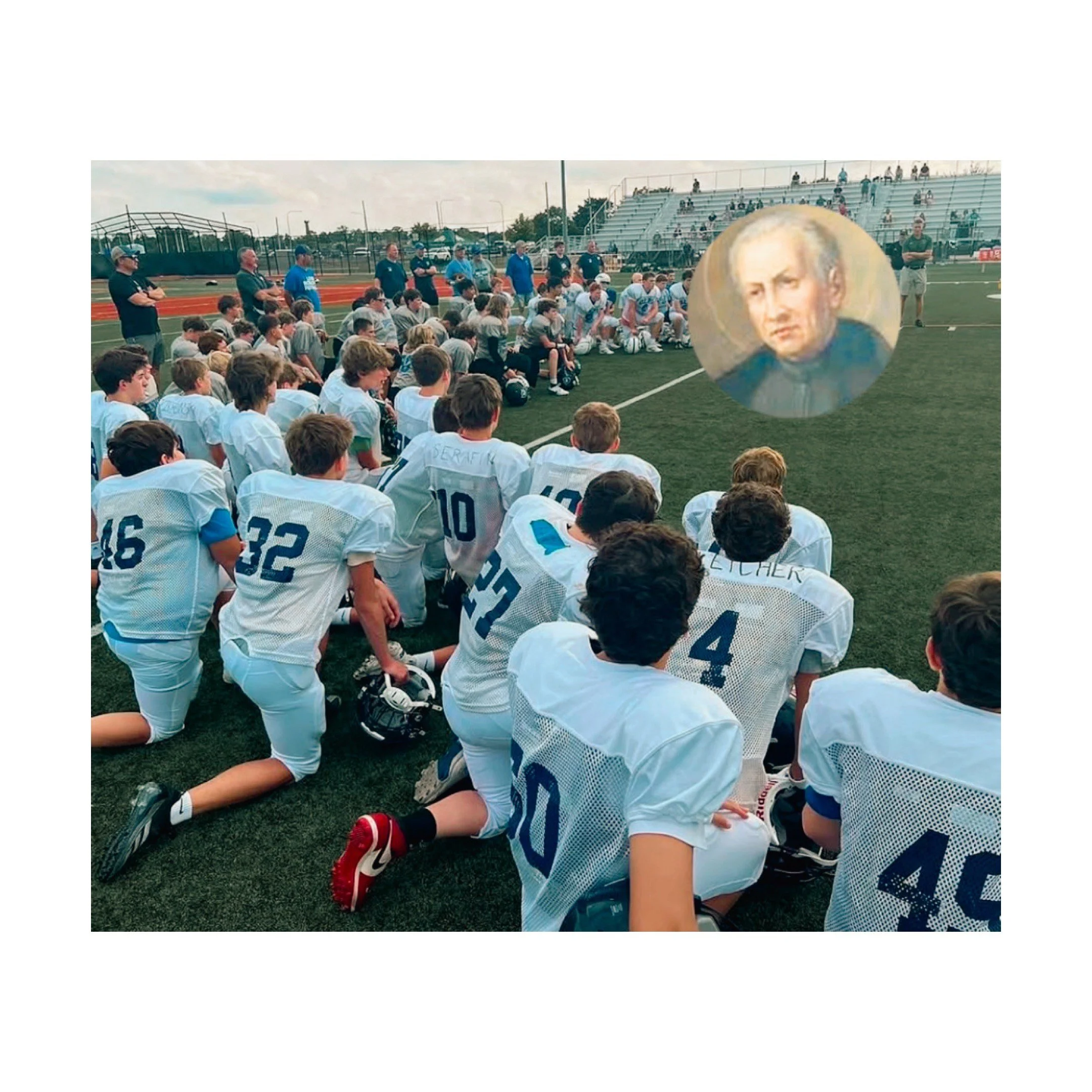 A youth football team kneeling on the field during a team huddle, with coaches and spectators in the background.