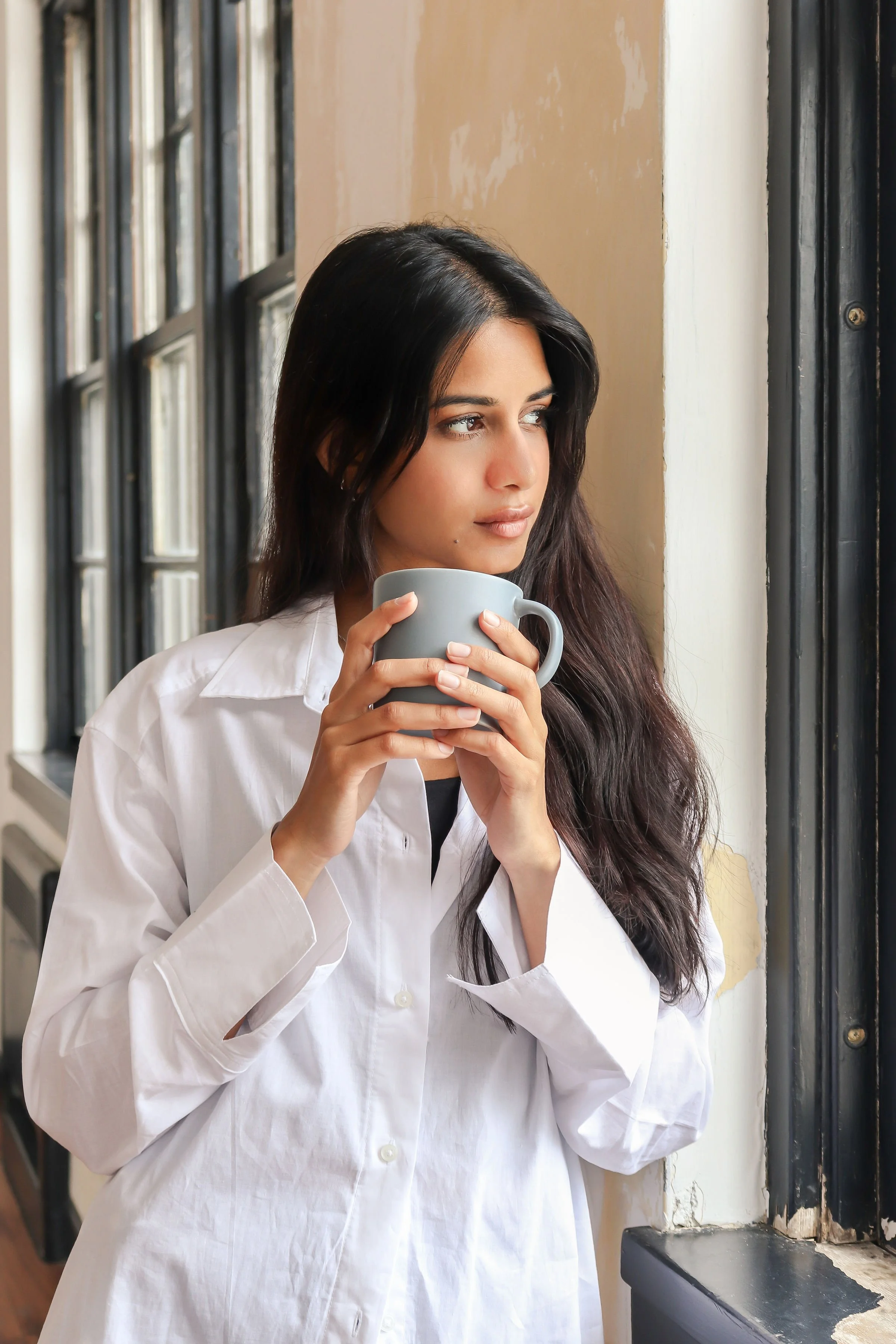 A young woman with long dark hair and light skin holding a grey mug, looking out a window with black metal frames and a distressed beige wall behind her.