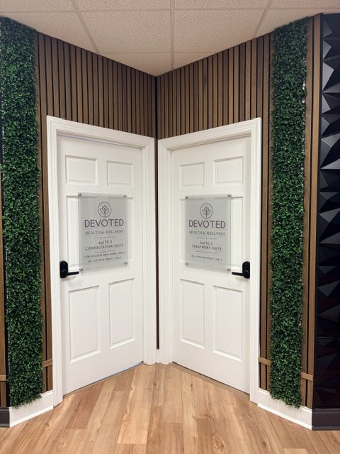 Corner of a room with two closed white doors with signs, surrounded by green artificial plants and brown wood paneling walls.