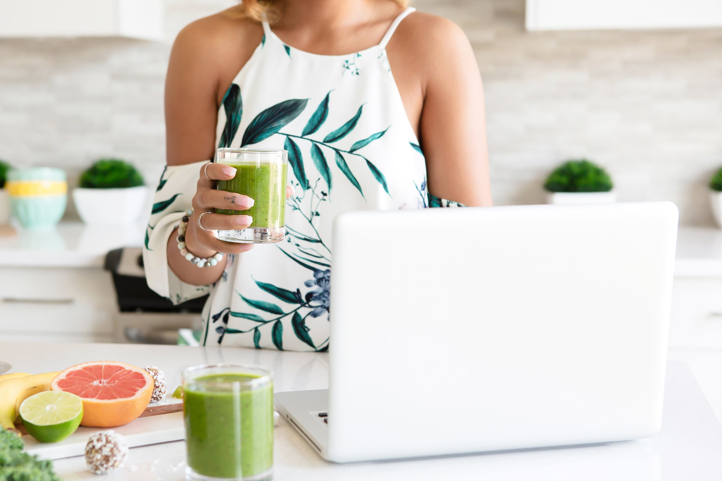 Woman holding a glass of green smoothie in a kitchen with fruits and a laptop on the counter.