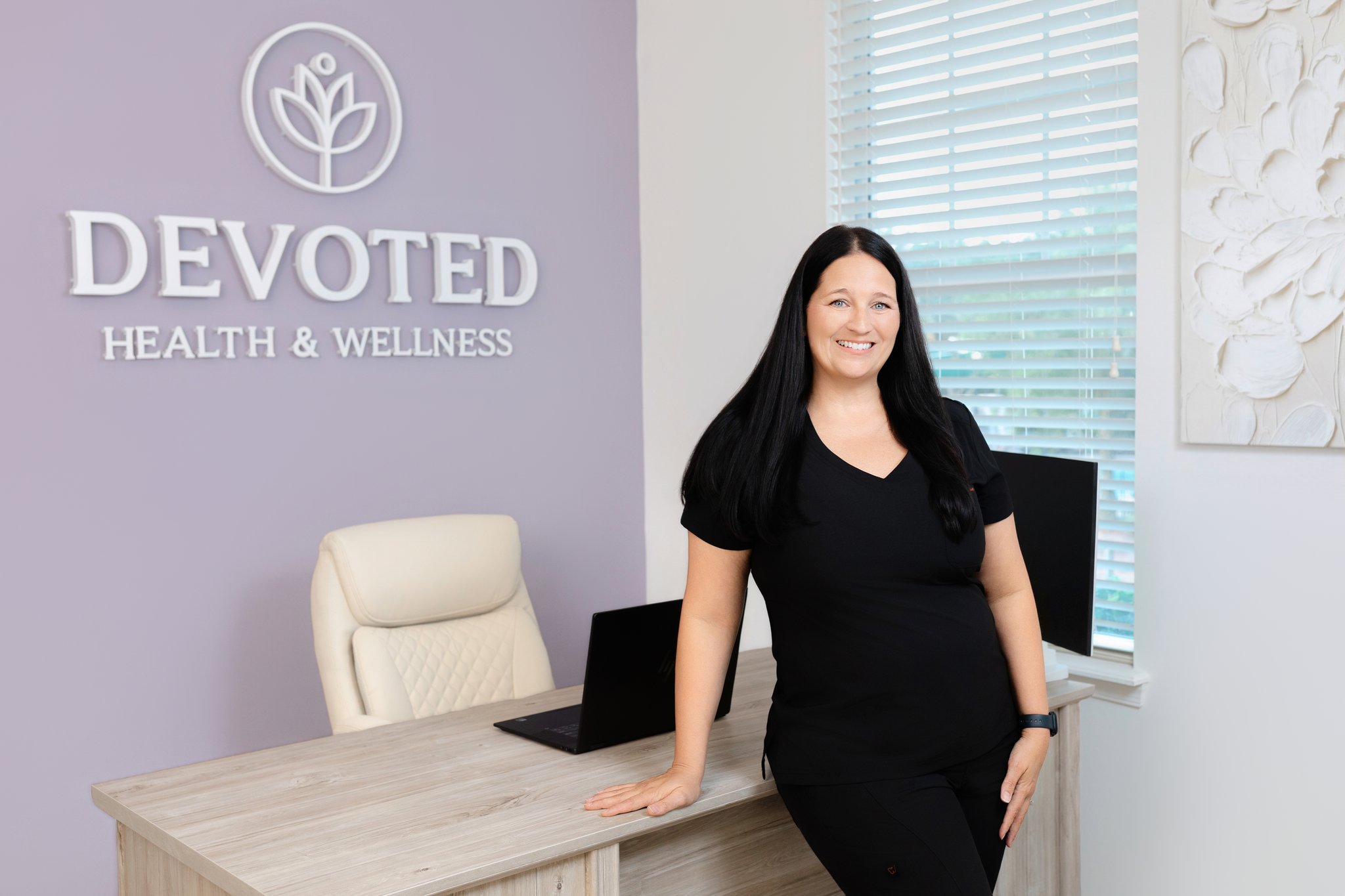A woman with long black hair and a black shirt leaning on a wooden desk with a laptop in a wellness clinic. The wall behind has a purple sign that reads 'Devoted Health & Wellness' with a logo of a stylized flower. The room has large windows with blinds and white textured wall art.