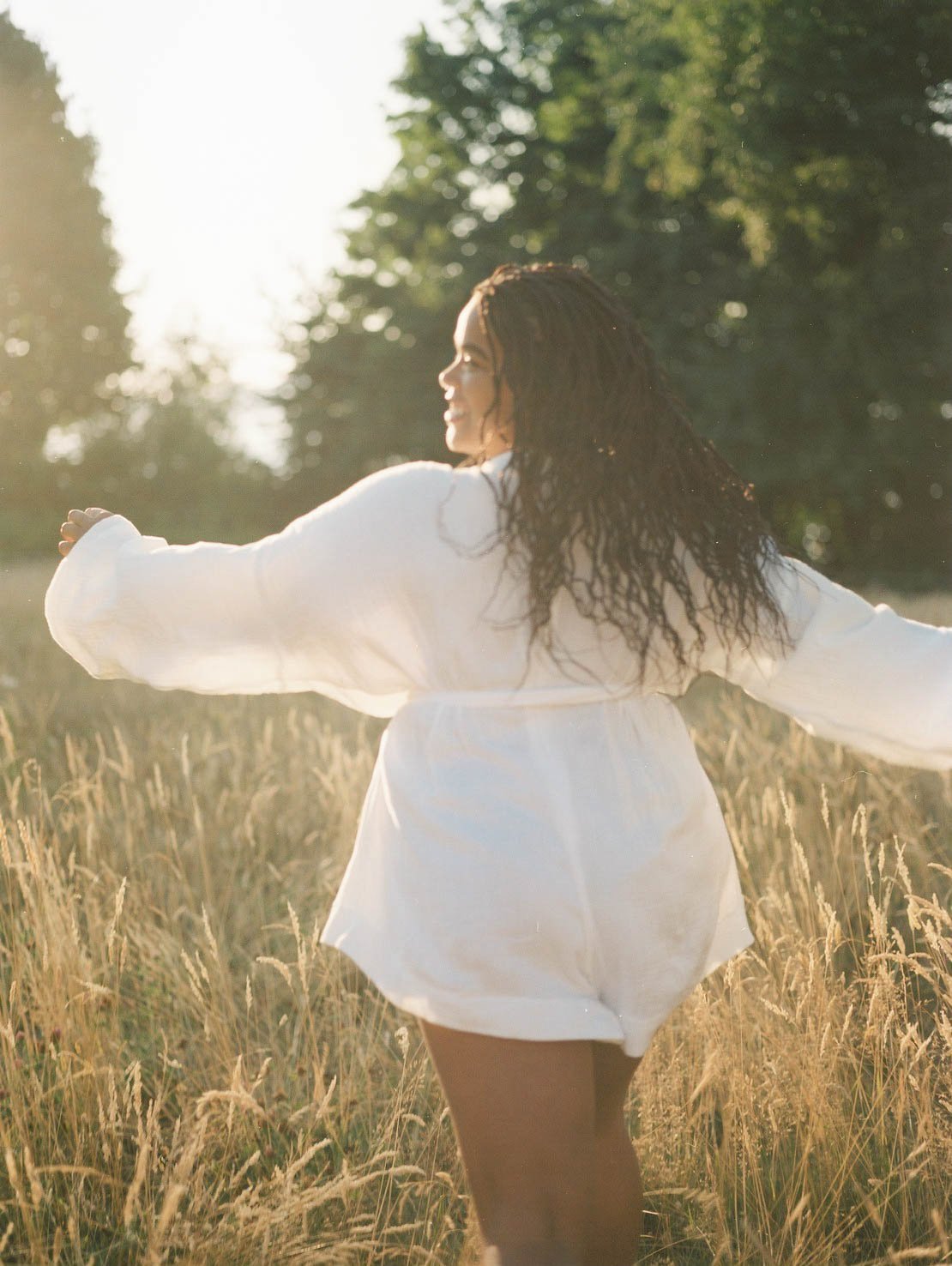 A woman with long curly hair wearing a white dress standing in a field with arms outstretched during sunset.