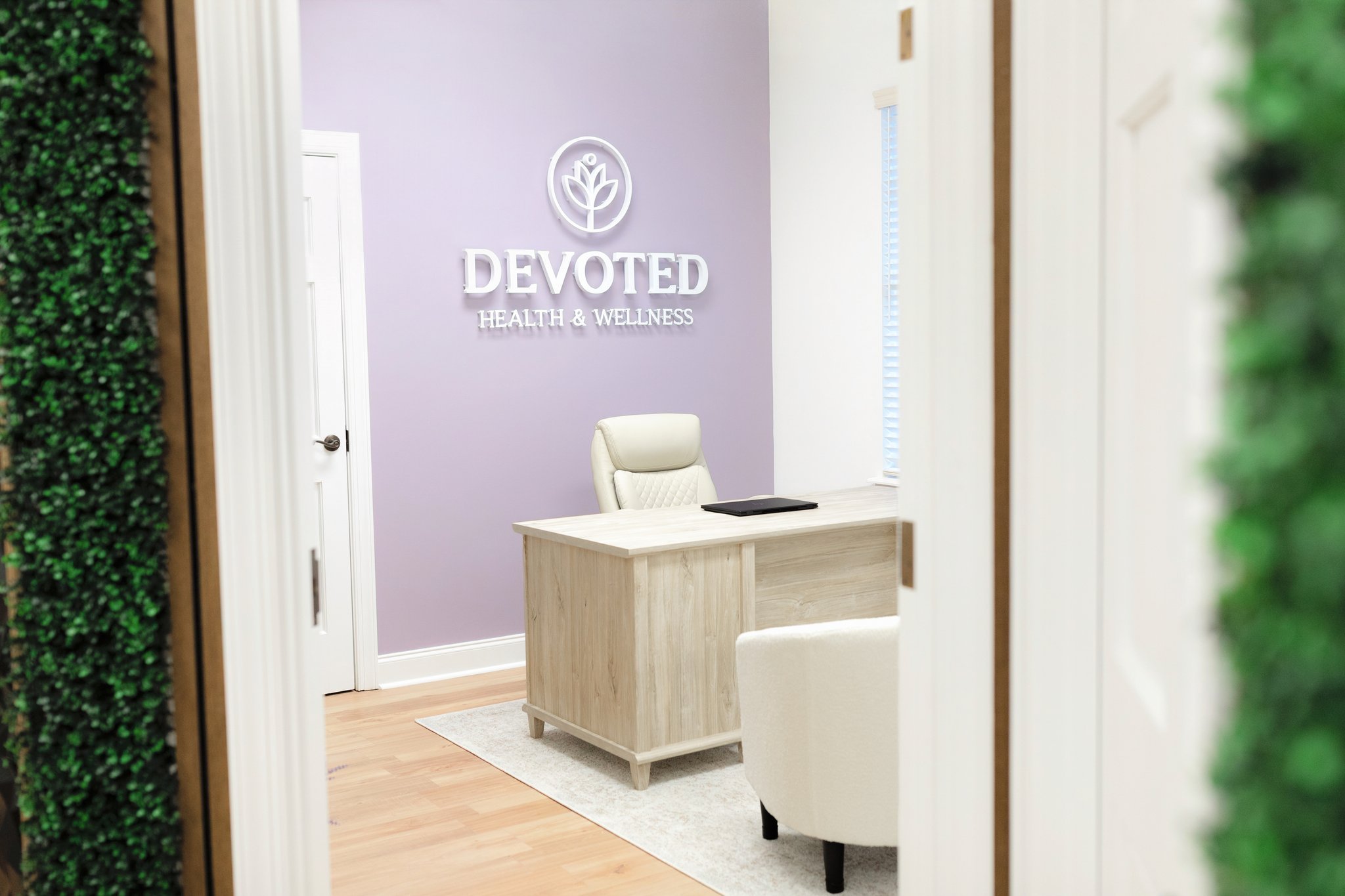 Reception area at Devoted Health & Wellness with a white desk, chair, purple accent wall, and a decorative sign featuring a flower logo.