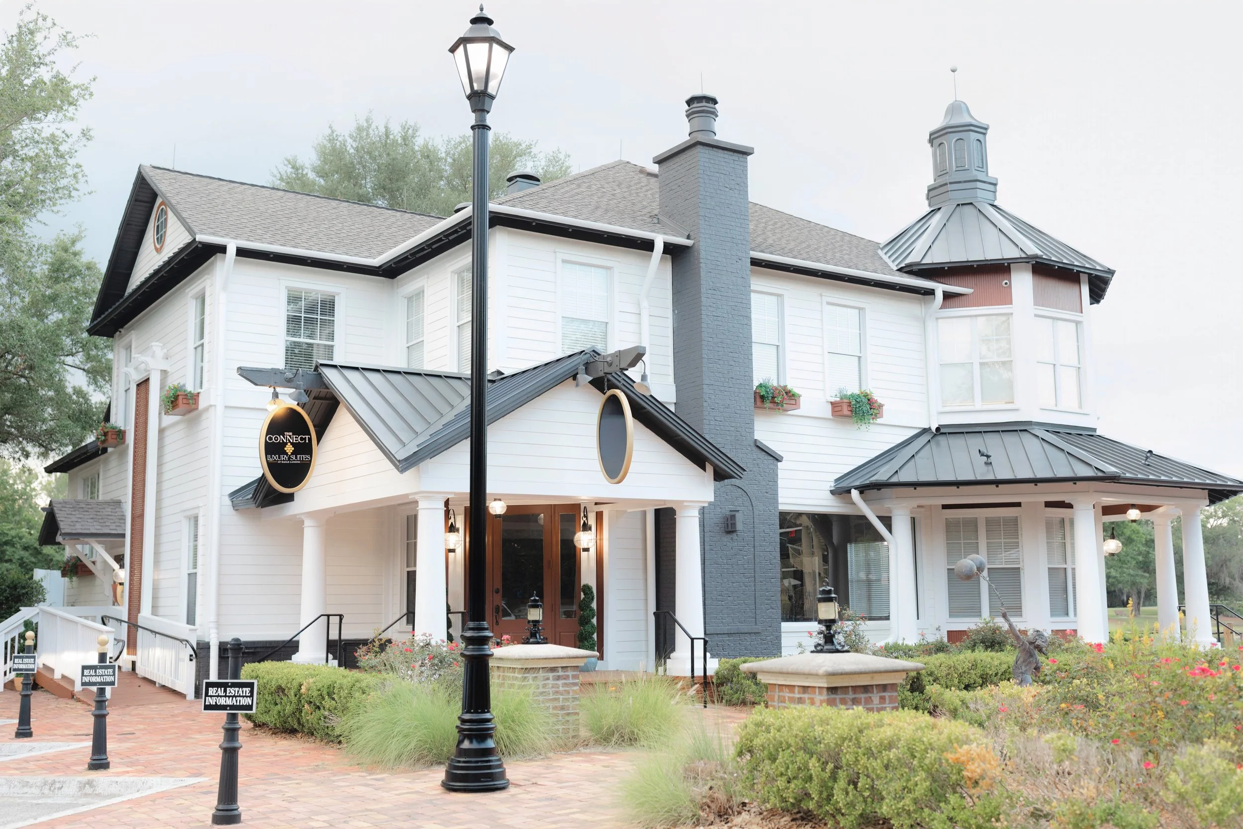 A large, multi-story house with white siding, black accents, and a gray roof. It has a prominent central chimney and a turret with a conical gray roof. The house features multiple windows with flower boxes. The front yard is landscaped with bushes, f