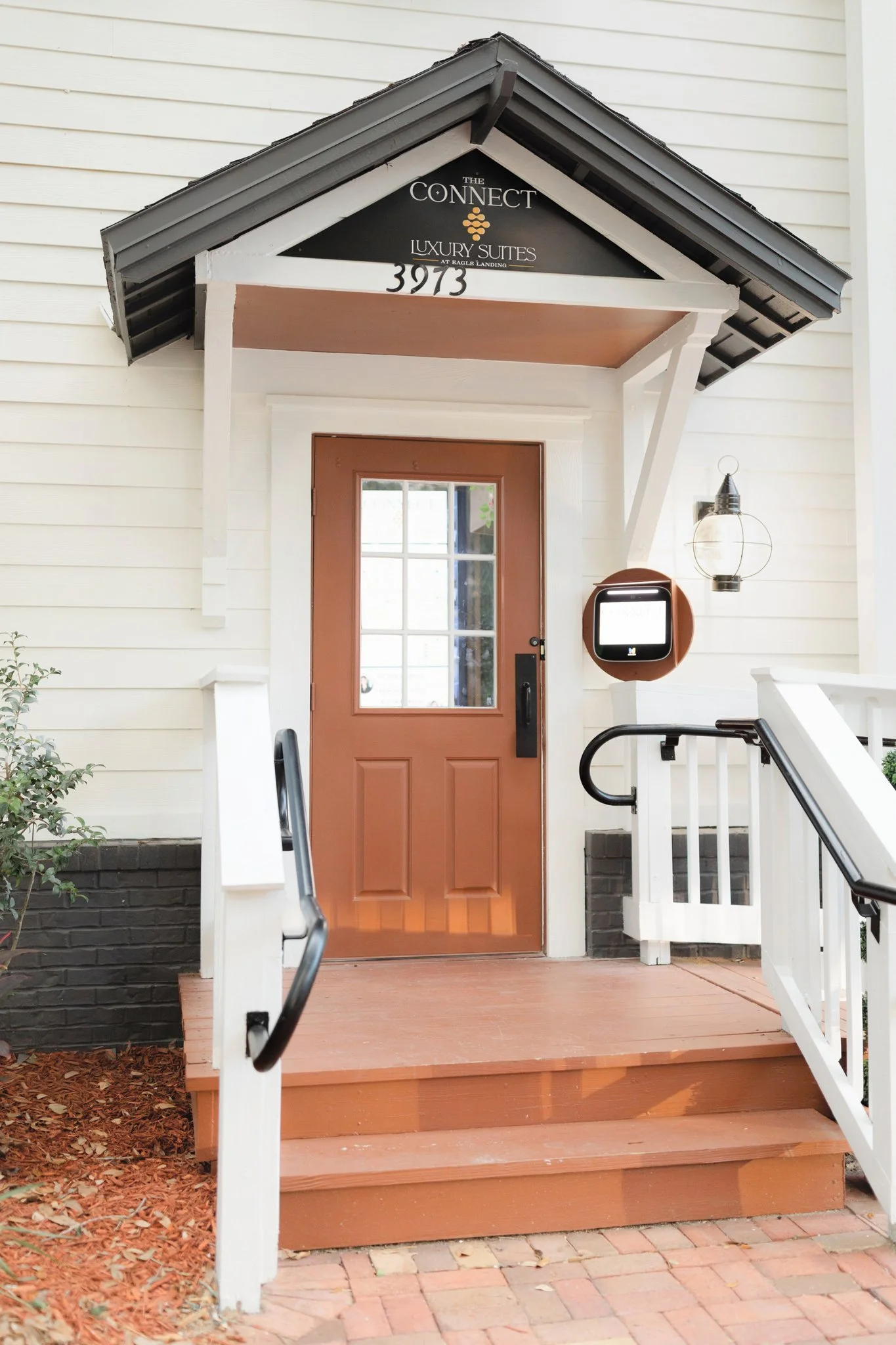 Front porch of a building with a brown door, white trim, and a black and white sign. There are steps with black railings and a porch light on the right side.