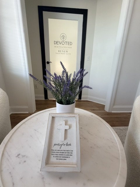 A round white marble table with a small prayer box and a white pot of purple lavender flowers on top, with a door in the background that has the words "Devoted Therapeutic Wellness" and "Renew Treatment Suite" on it.