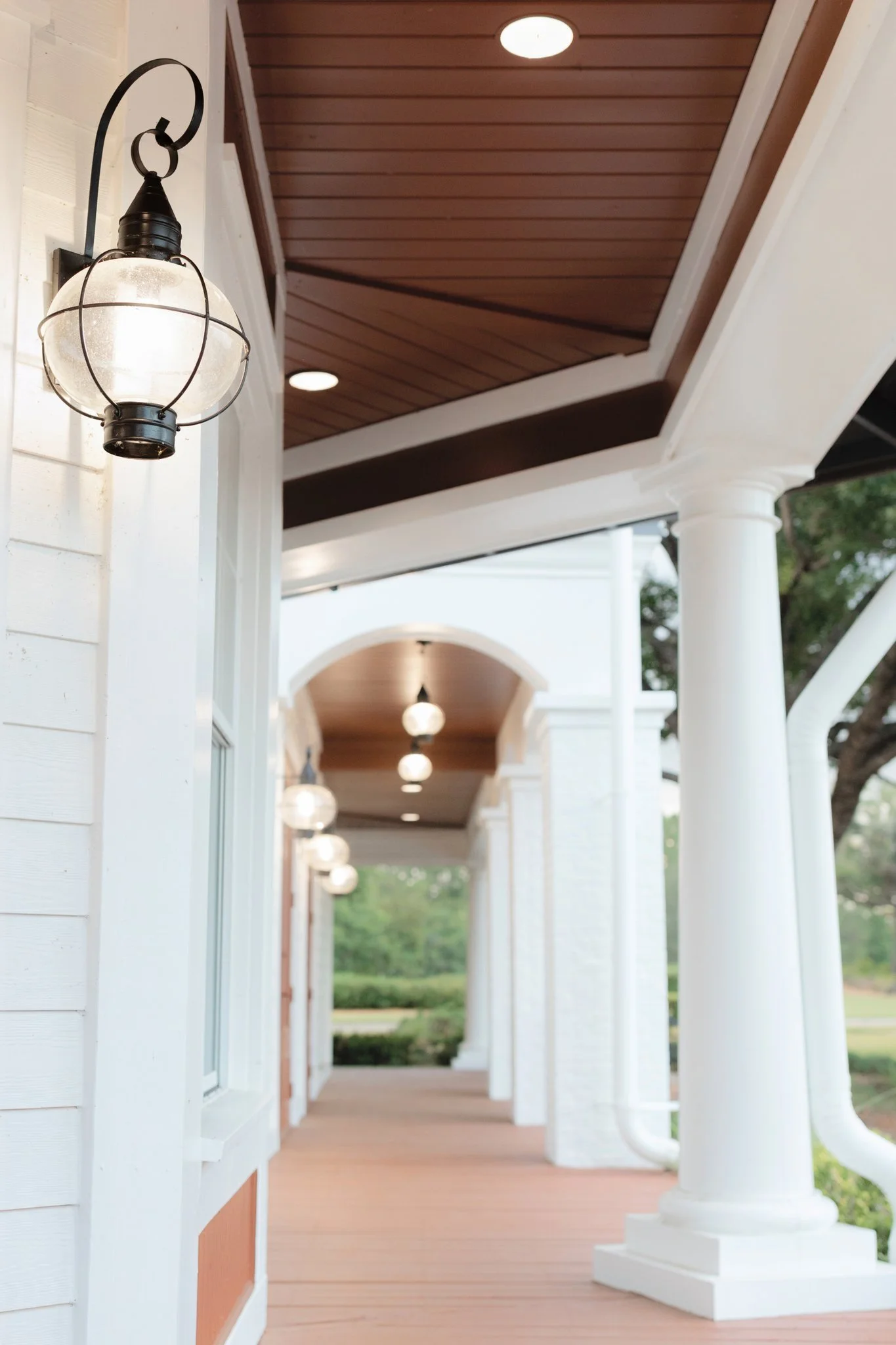 A porch with white columns, a brown wooden ceiling, hanging round light fixtures, and a view of greenery in the background.