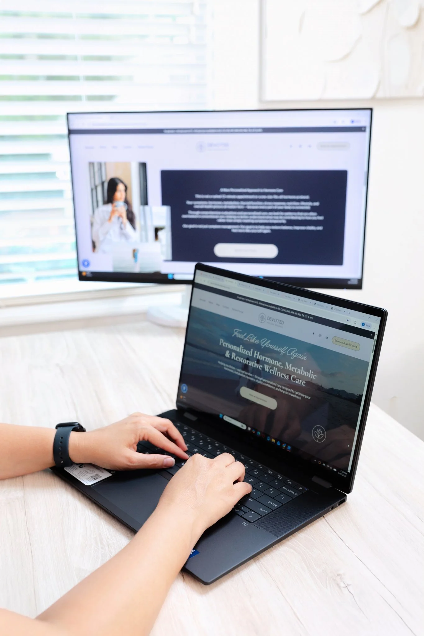 A person using a laptop computer on a light wood desk, with a larger monitor in the background displaying a website with a woman holding a phone.