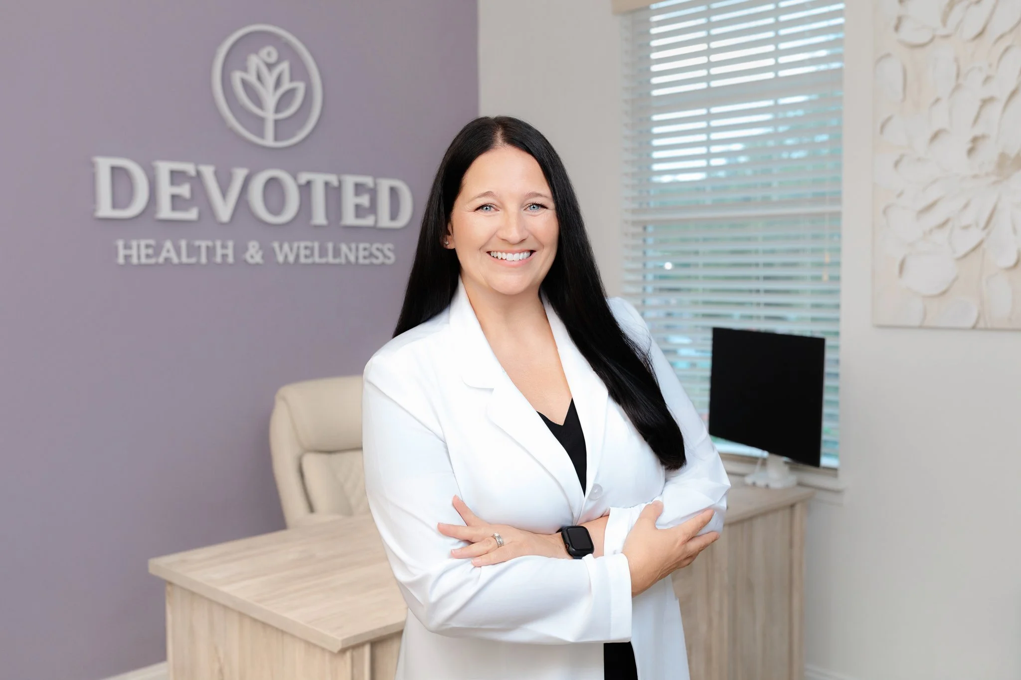A woman with long dark hair, dressed in a white blazer, stands with her arms crossed, smiling in an office environment. Behind her, there is a sign on the wall that reads 'Devoted Health & Wellness' with a logo of a stylized plant. The office has a window with blinds and modern decor.