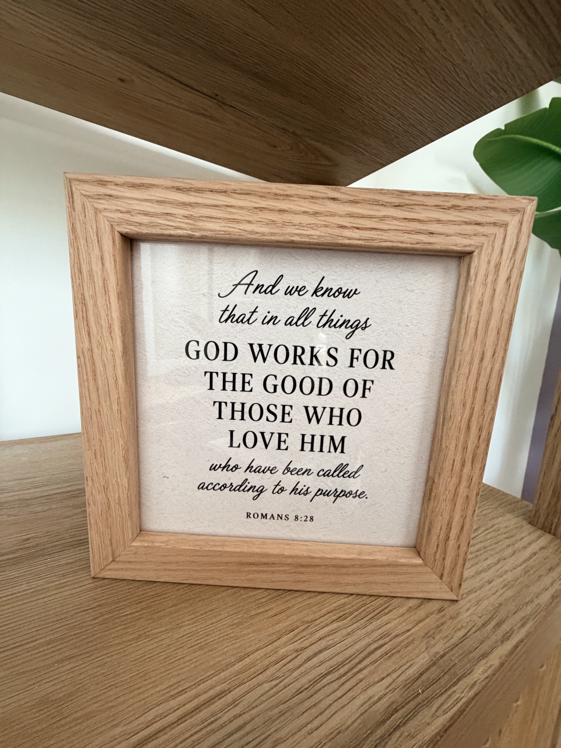 A framed quote on a wooden surface reads: "And we know that in all things GOD WORKS FOR THE GOOD OF THOSE WHO LOVE HIM" followed by a verse reference "ROMANS 8:28". The frame is light wood, and there is a green leaf partially visible on the right sid