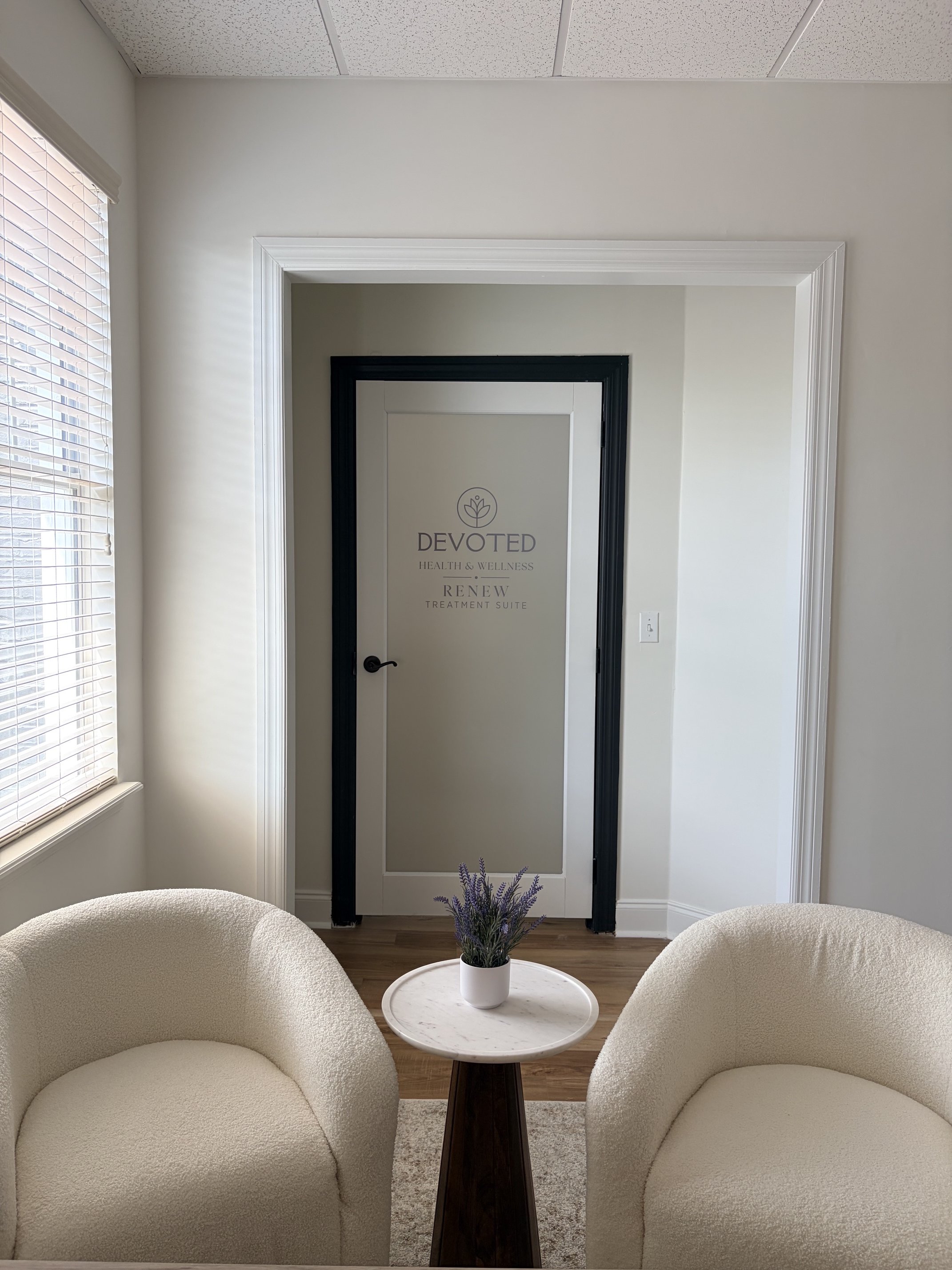 Waiting area with two cream-colored, rounded armchairs on either side of a small white marble-top table with a potted purple lavender plant. Behind, a doorway with black and white trim leads to a treatment room with the words 'Devoted Health & Wellne