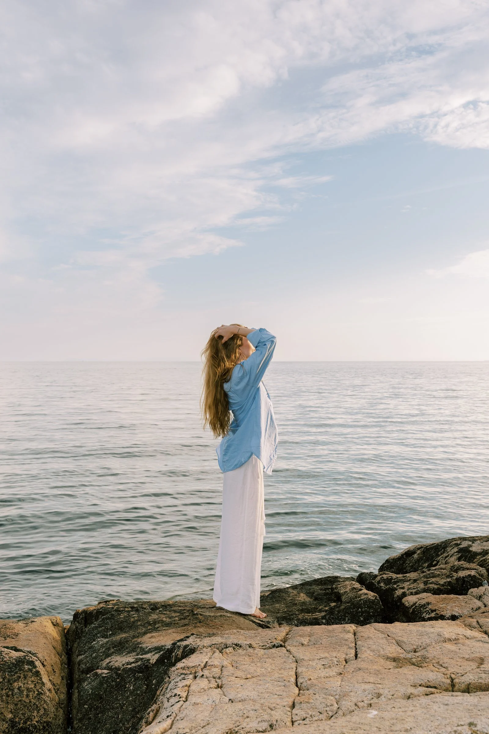 A woman with long wavy hair wearing a blue shirt and white skirt standing on rocks near the sea, holding her head with both hands, with a cloudy sky overhead.