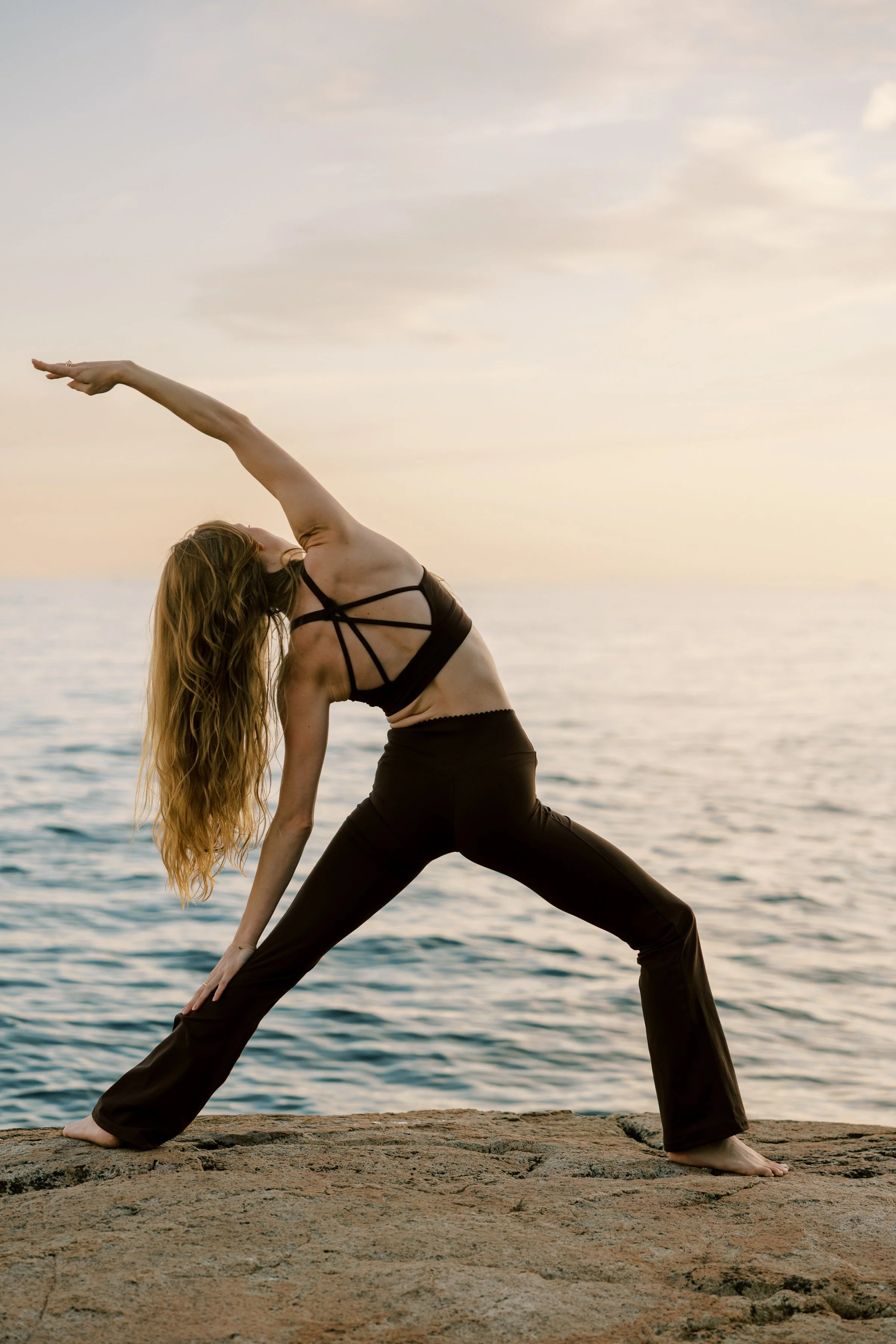 A woman with long hair in yoga pose on rocks by the water during sunset.