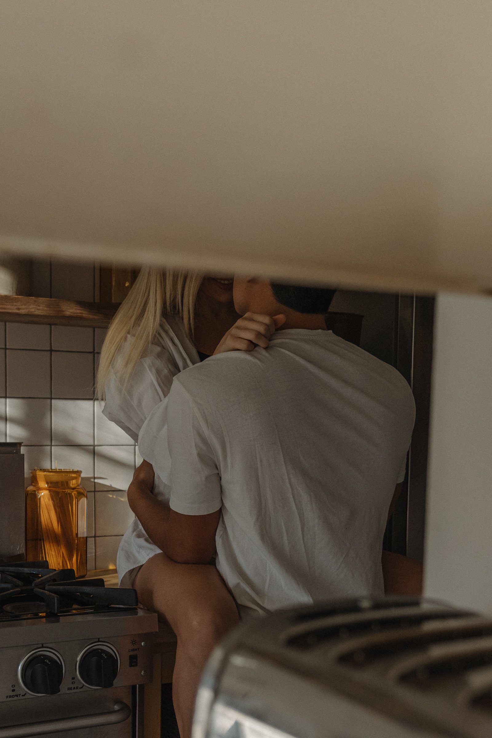 A couple kissing in a kitchen, seen through a window, with the woman having blonde hair and wearing a white shirt, and the man in a white shirt.