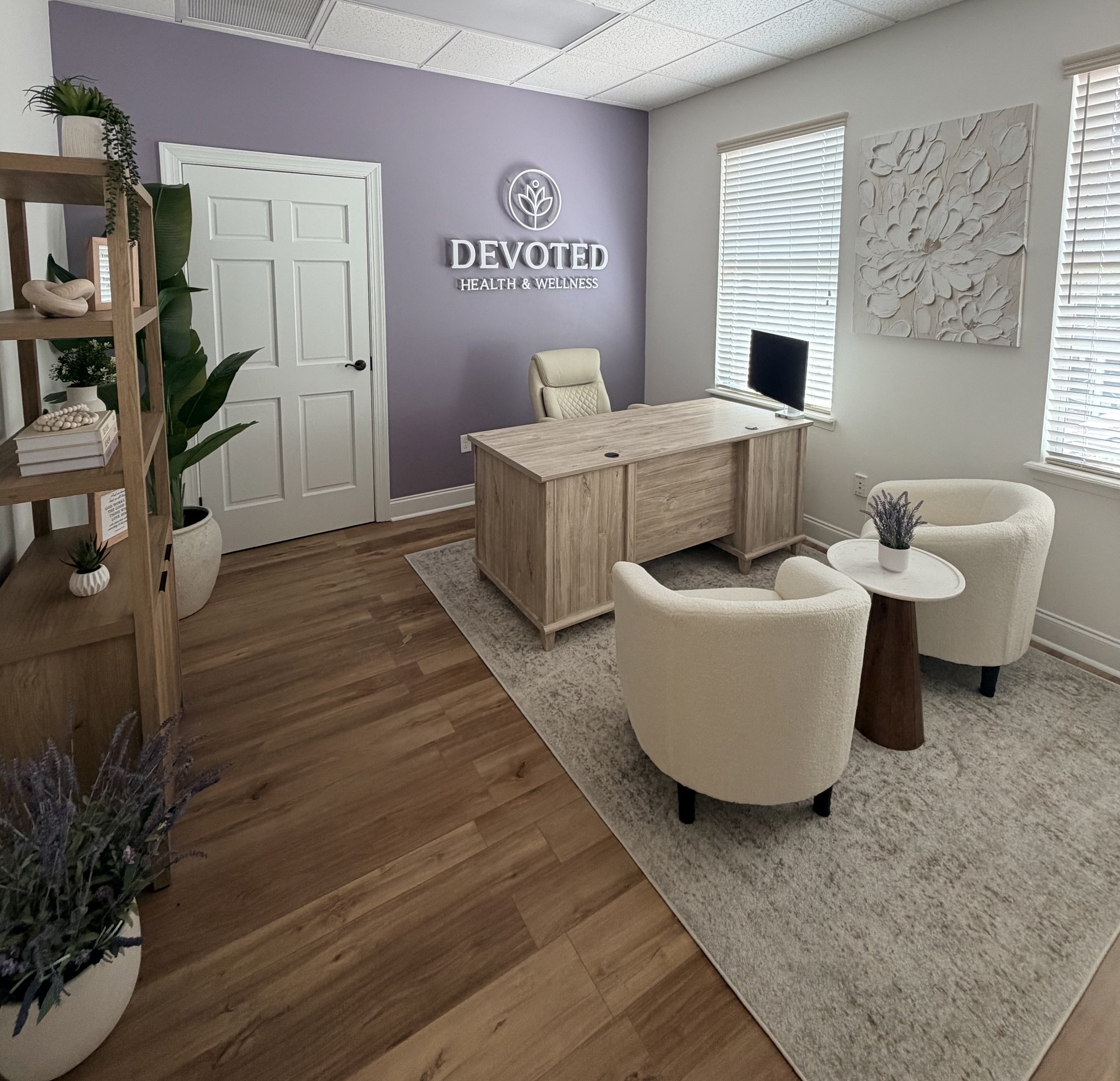 Reception area with a sign reading 'Devoted Health & Wellness' on a lavender wall, a desk with a computer, a white armchair, decorative plants, artwork, and large windows with blinds.