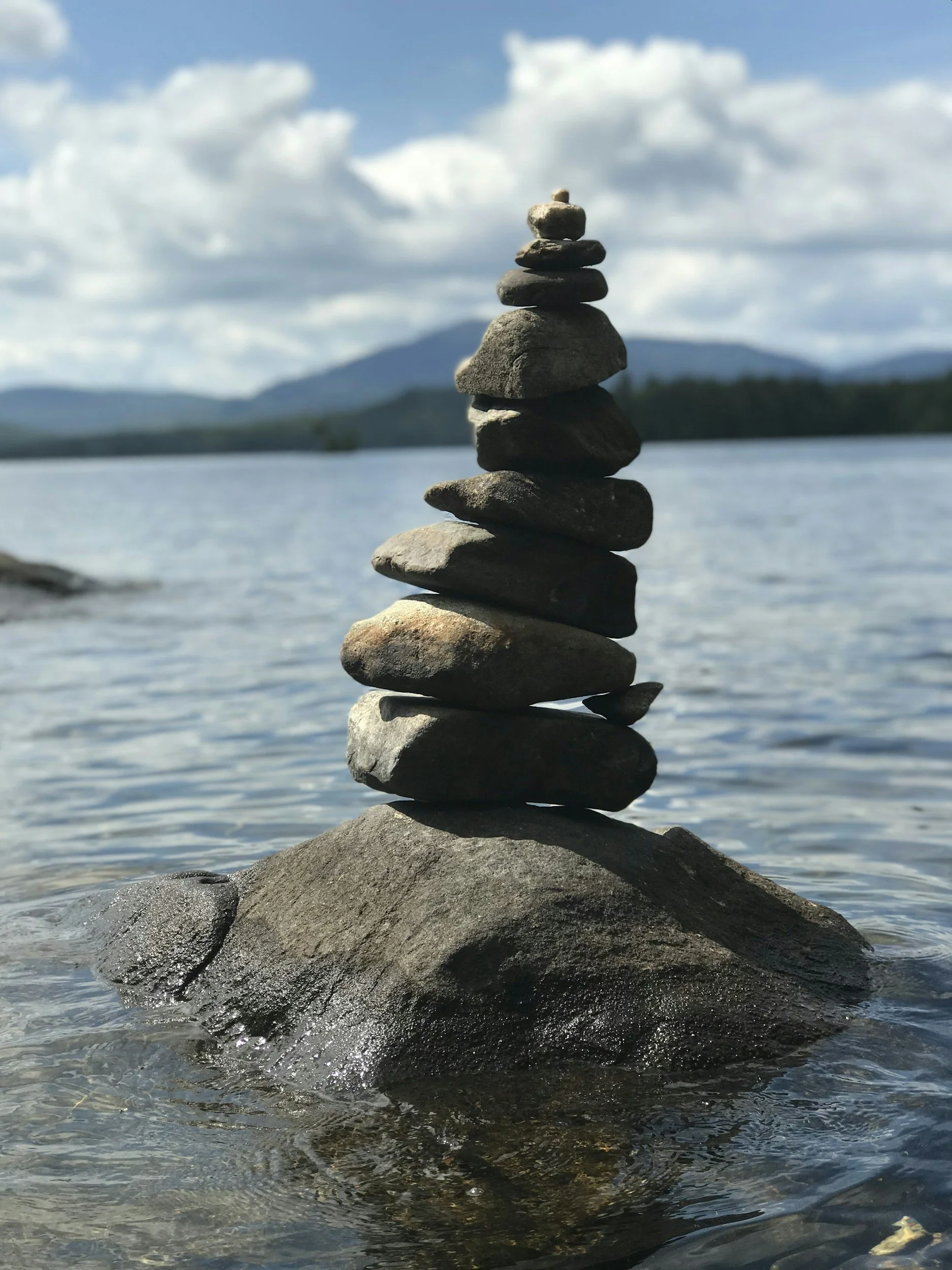 Stack of balanced stones on a rock in a calm lake with mountains and a cloudy sky in the background.