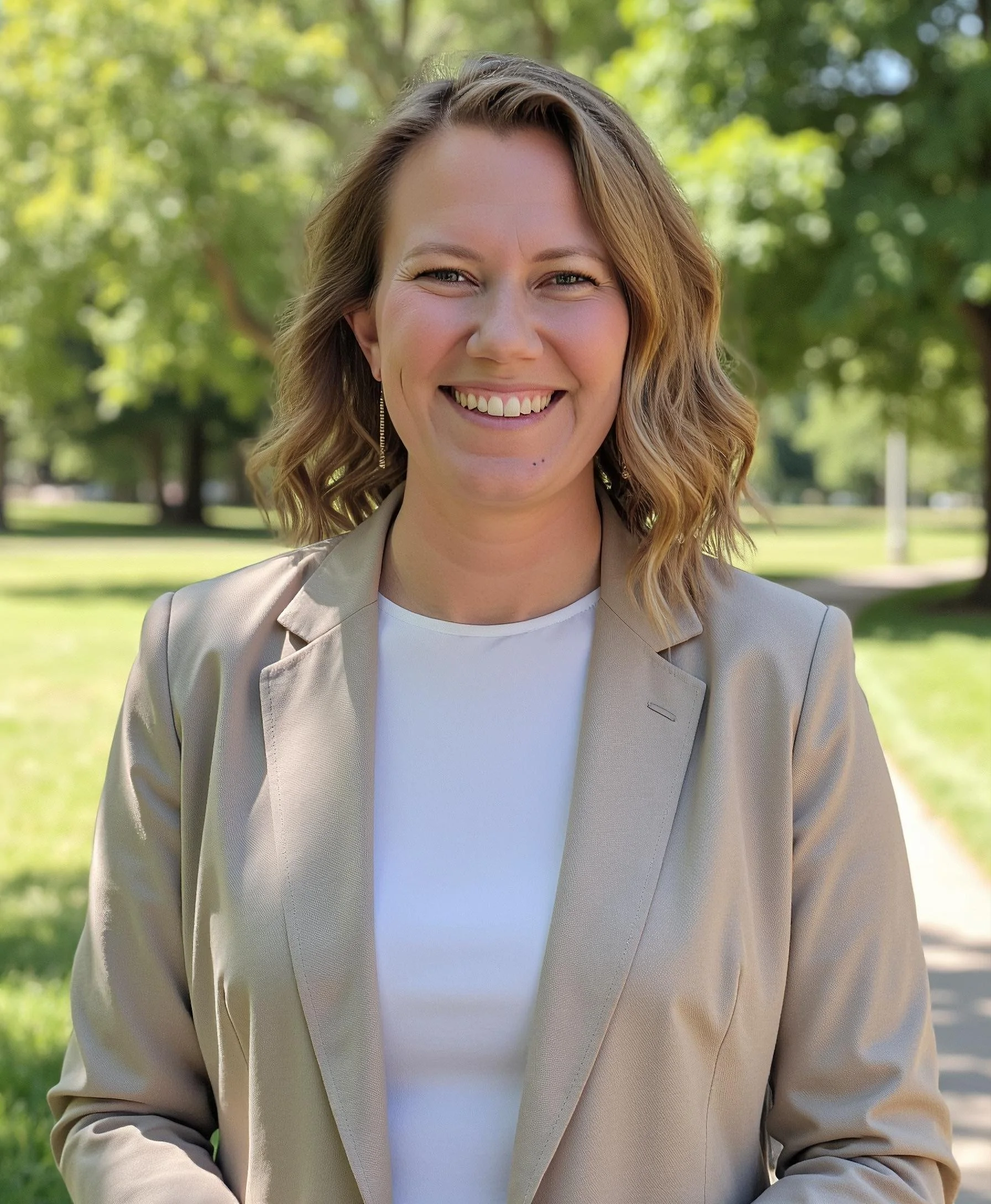 Woman smiling in a beige blazer and white shirt, standing outdoors with trees and grass in the background.