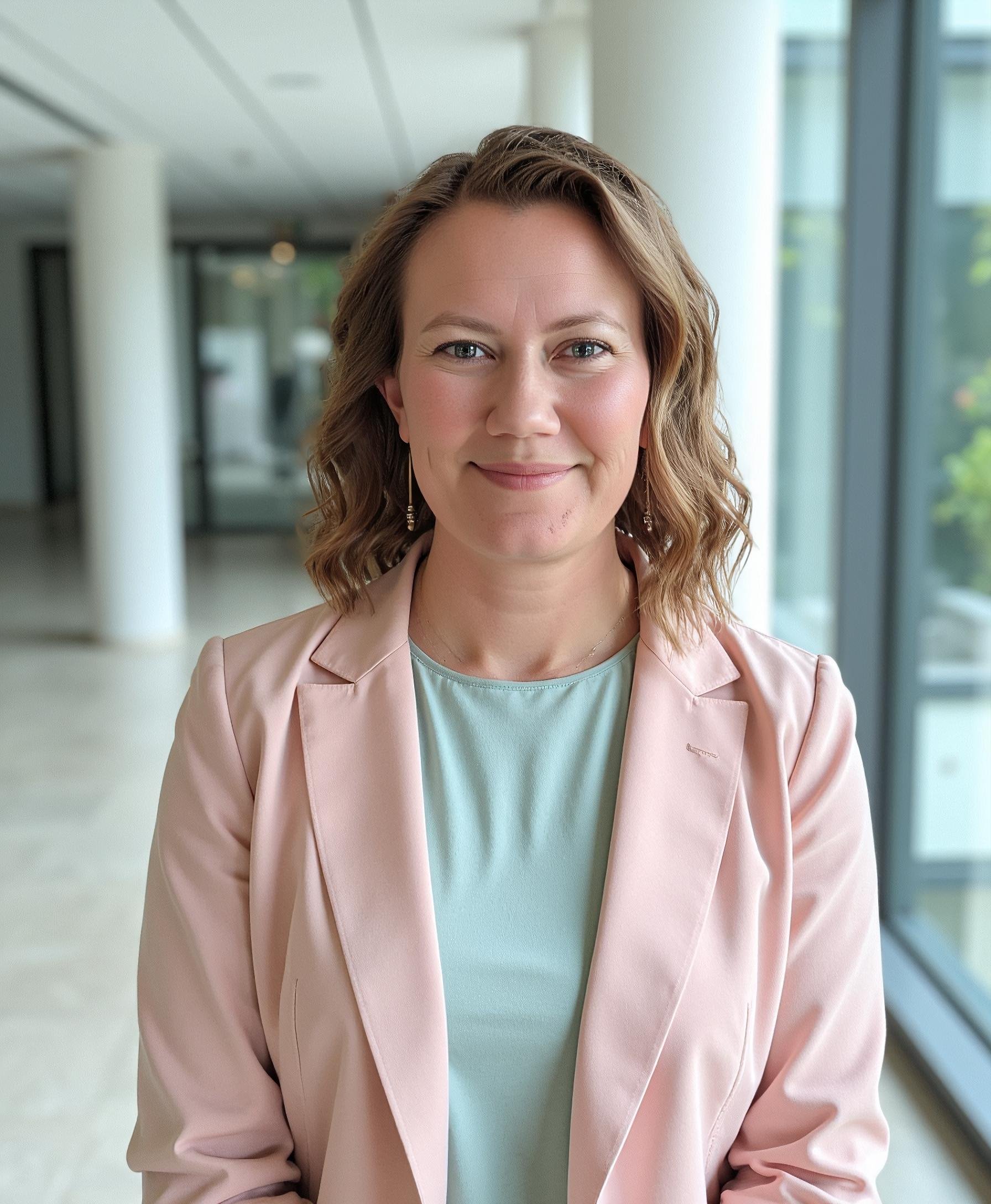 Woman smiling in a professional setting, wearing a pink blazer and light green top, standing in a hallway with large windows.