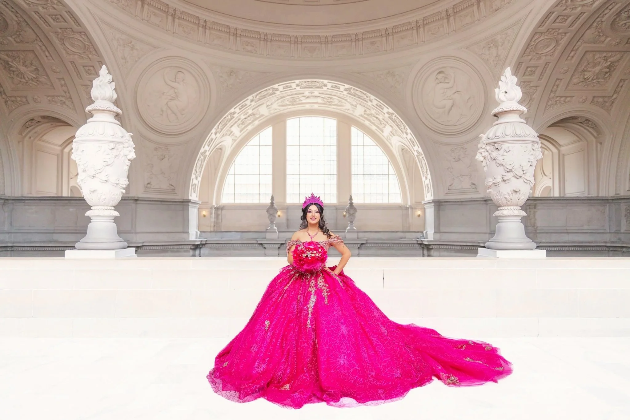 Quinceañera in a bright pink ball gown posed beneath an architectural arch at San Francisco City Hall, with the gown flowing across the floor.