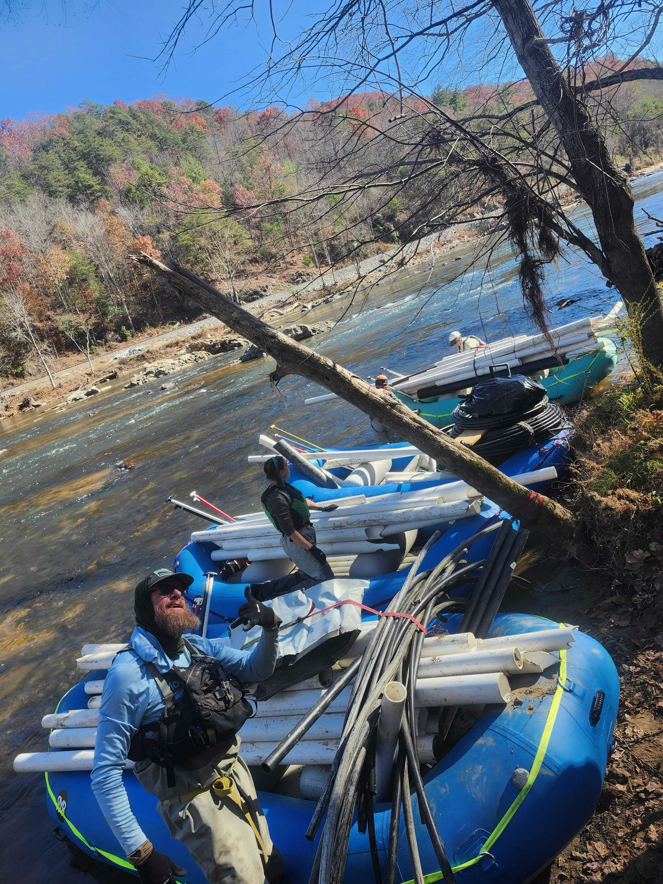 Two men preparing a raft with PVC pipes and other supplies on the rocky shore of a river, with trees and a hillside in the background, under a clear blue sky.