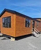 Wooden tiny house with stairs, set on a gravel lot under a blue sky.
