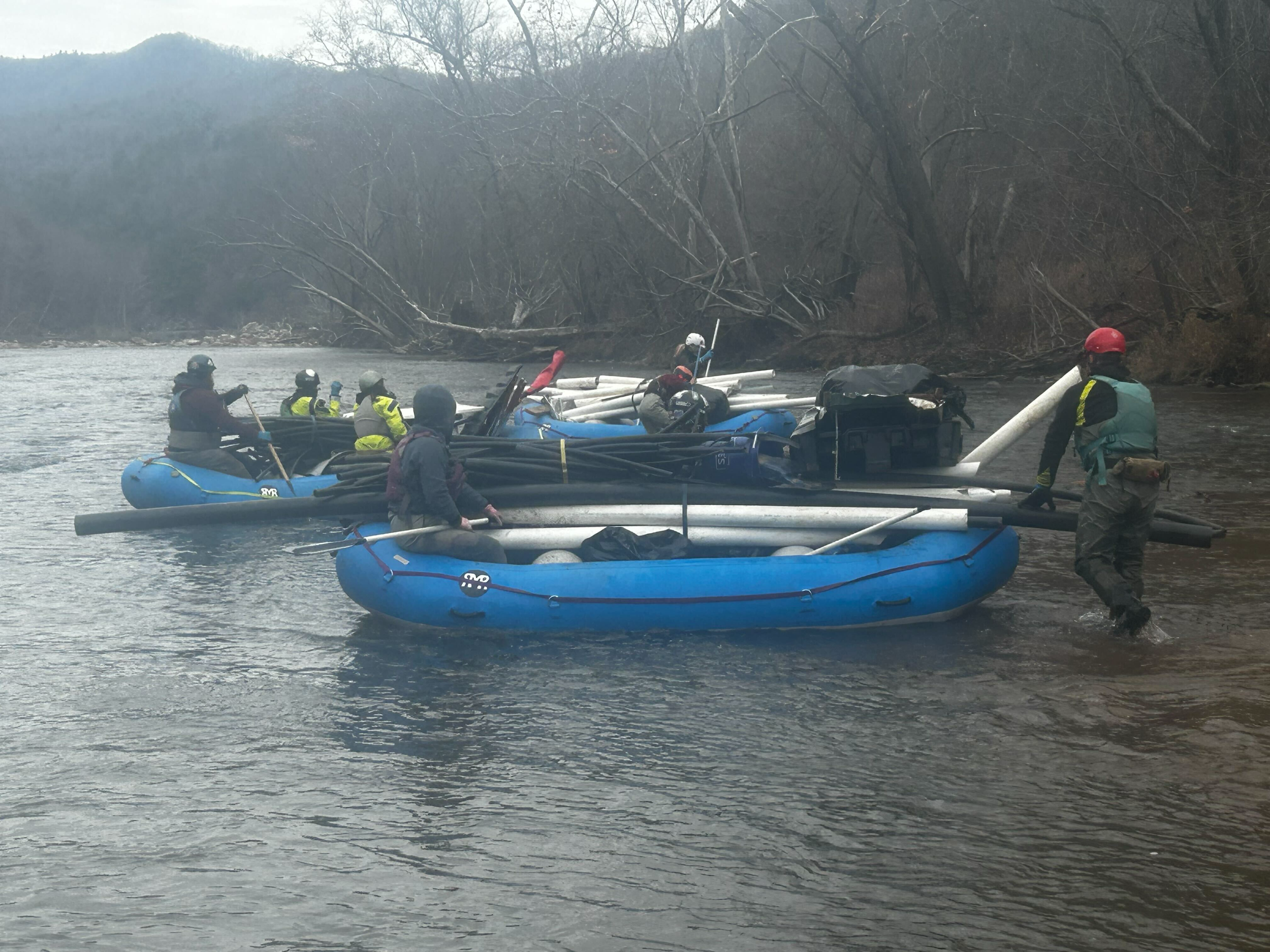A group of people in kayaks on a river, wearing helmets and life jackets, with fallen trees and bare trees in the background.