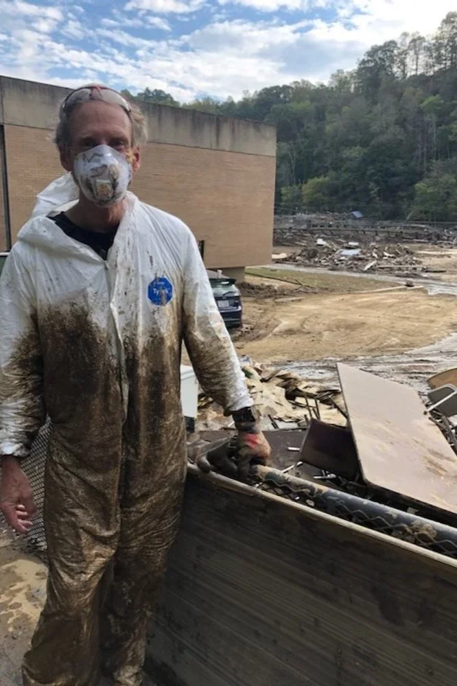 A man wearing a dirty white jumpsuit and a face mask stands outside near a building, holding a power tool. The area around him is muddy and debris is scattered across the ground, suggesting recent flooding or natural disaster.
